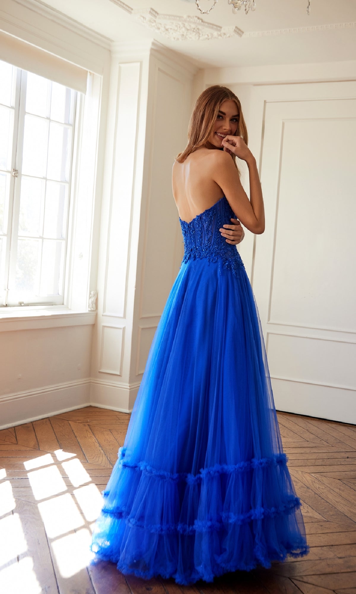 A woman smiles in a bright room, wearing the Juliet JT2561S strapless long prom ball gown with an embroidered-lace bodice and tulle skirt; the elegant blue dress fits her beautifully as she faces slightly away.