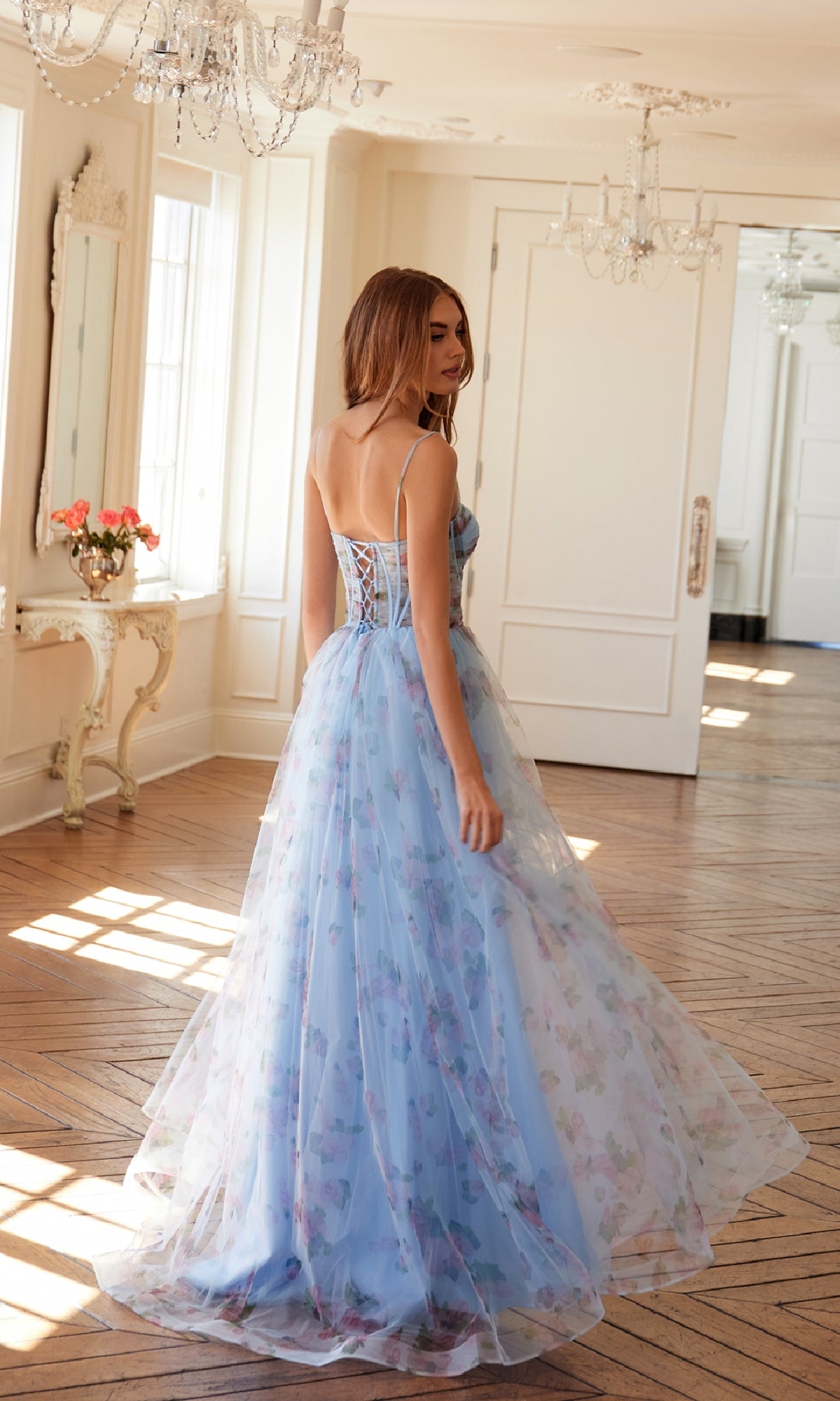 A woman wears the Strapless Blue Print Prom Gown: Juliet JT2562S, standing in an elegant, sunlit room with chandeliers and wooden floors.