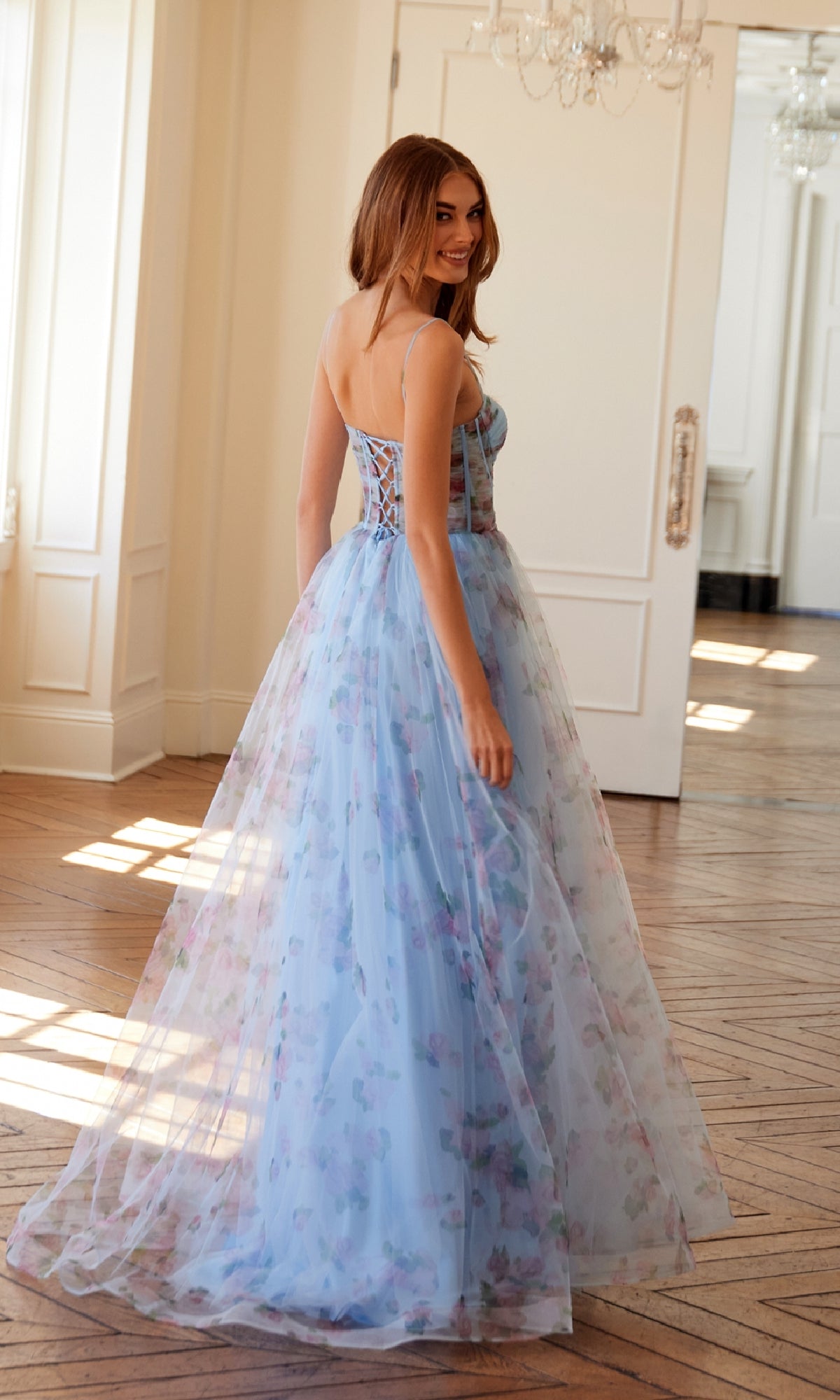 A woman in the Strapless Blue Print Prom Gown: Juliet JT2562S stands on wooden flooring indoors, smiling over her shoulder. The gown features a floral print and a lace-up corset bodice.