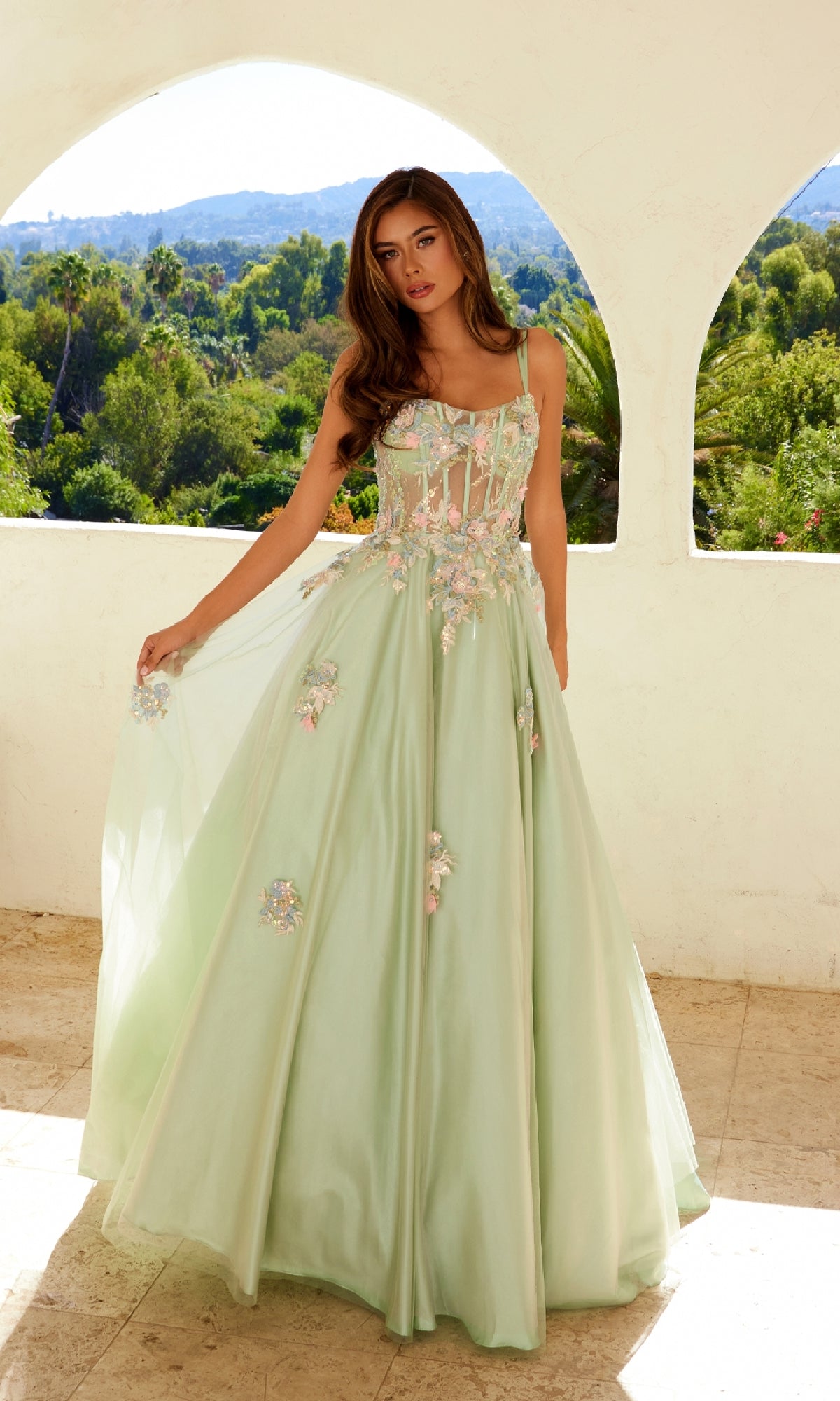 A woman in front of arched windows wears the Juliet JT2565U Long Prom Dress, featuring lace embroidery, floral appliqués, and a sheer corset bodice as she holds out the skirt. Greenery and hills can be seen in the background.
