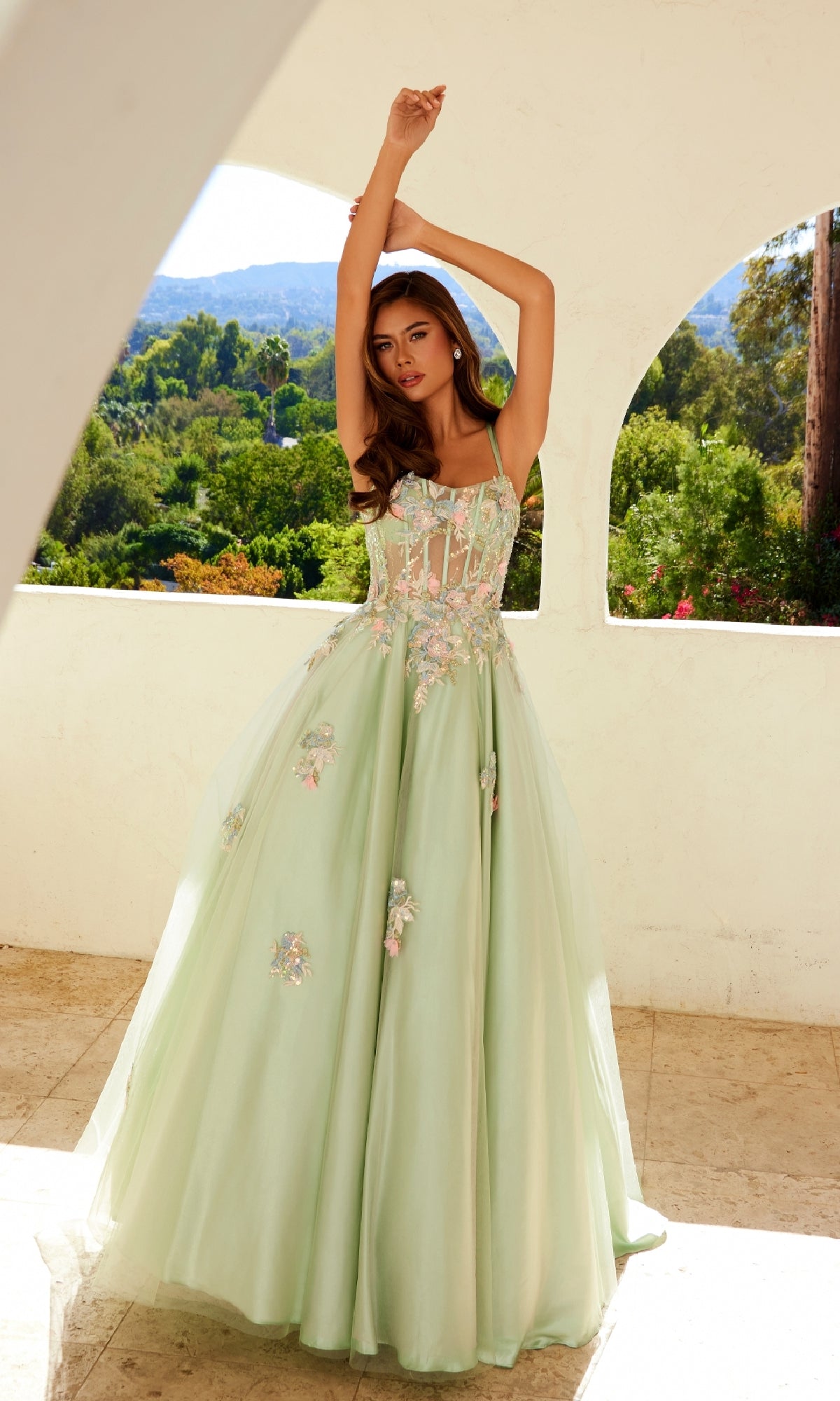 A woman poses with arms raised on a sunlit terrace, wearing the Juliet JT2565U Long Prom Dress—a light green gown with lace embroidery and a sheer corset bodice, surrounded by greenery and hills in the background.
