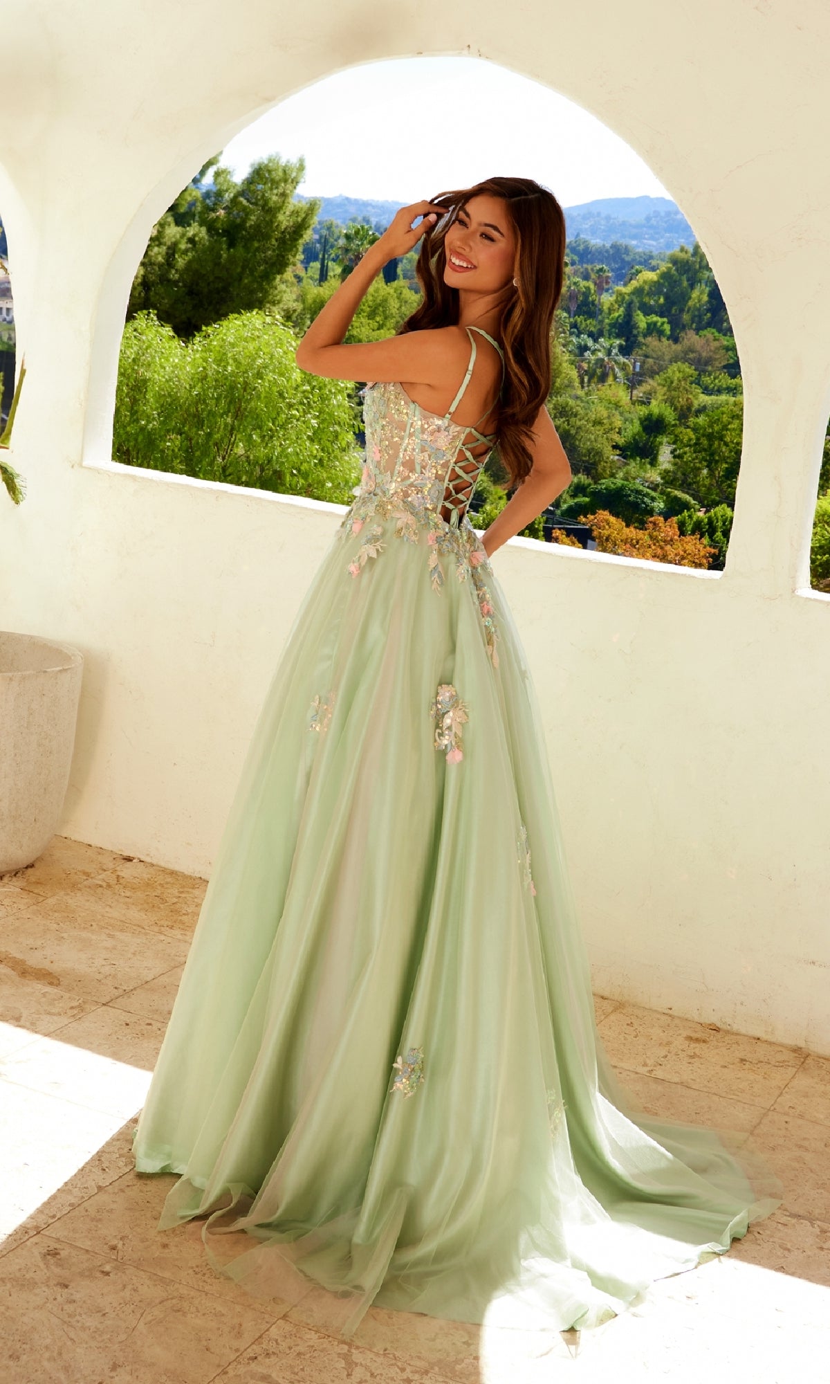A woman smiles over her shoulder on a patio, wearing the Juliet JT2565U long prom dress—a light green, floor-length ball gown featuring lace-embroidered floral details and a sheer corset bodice. Trees and hills fill the background.