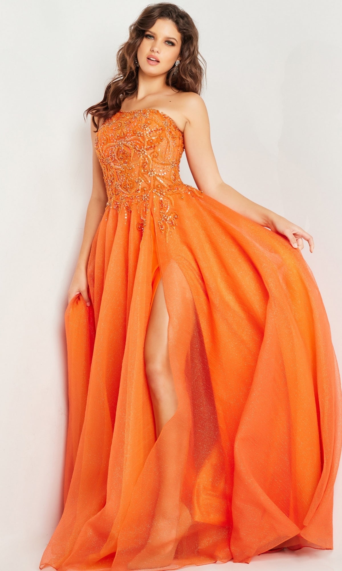 A woman poses in the Jovani One-Shoulder Long Prom Ball Gown 25688, featuring a vibrant orange hue, beaded bodice detailing, and a high slit in the flowing skirt.
