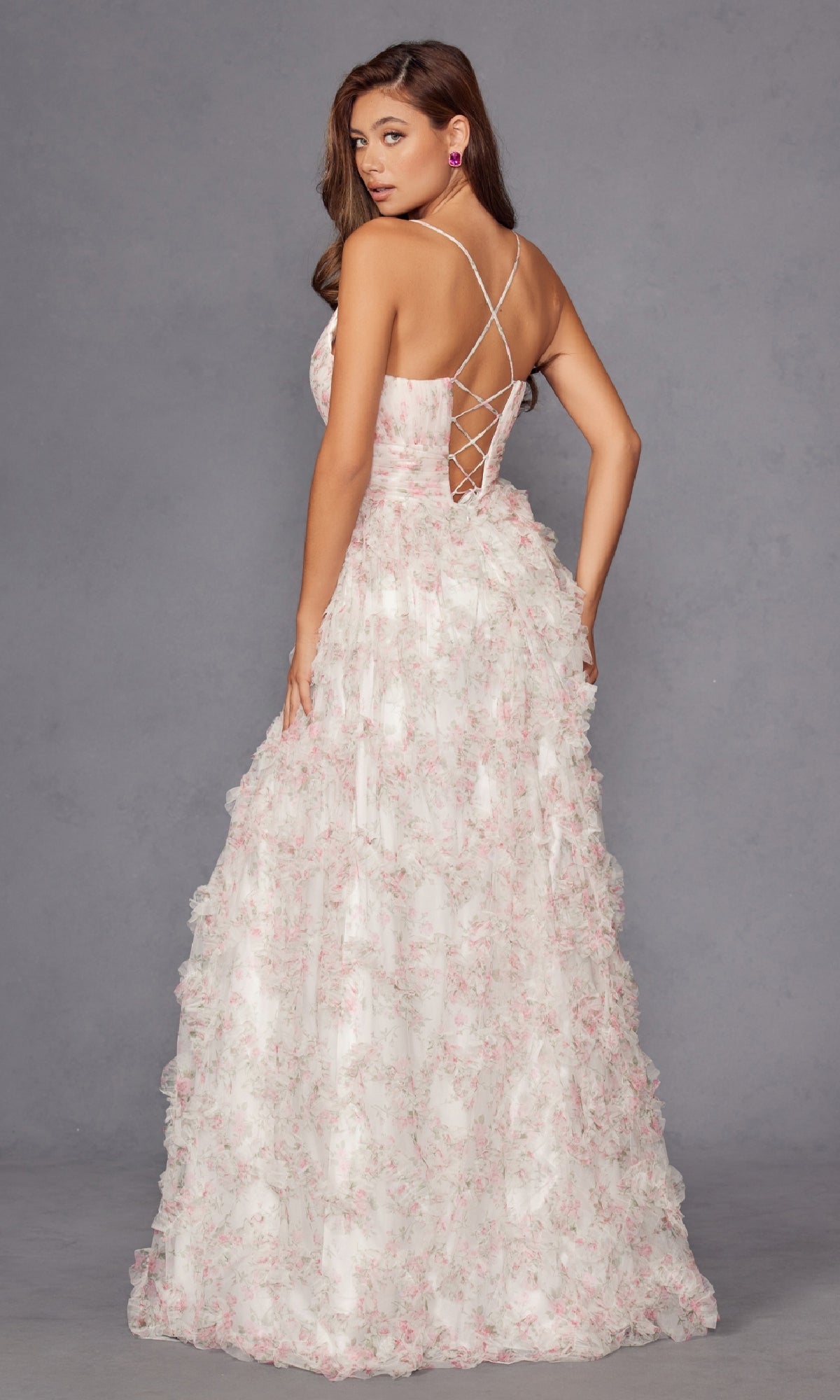 A woman models the Pink Floral Prom Dress: Juliet JT2572U, featuring a textured gown and lace-up back, standing on a plain grey backdrop, facing away from the camera while glancing over her shoulder.