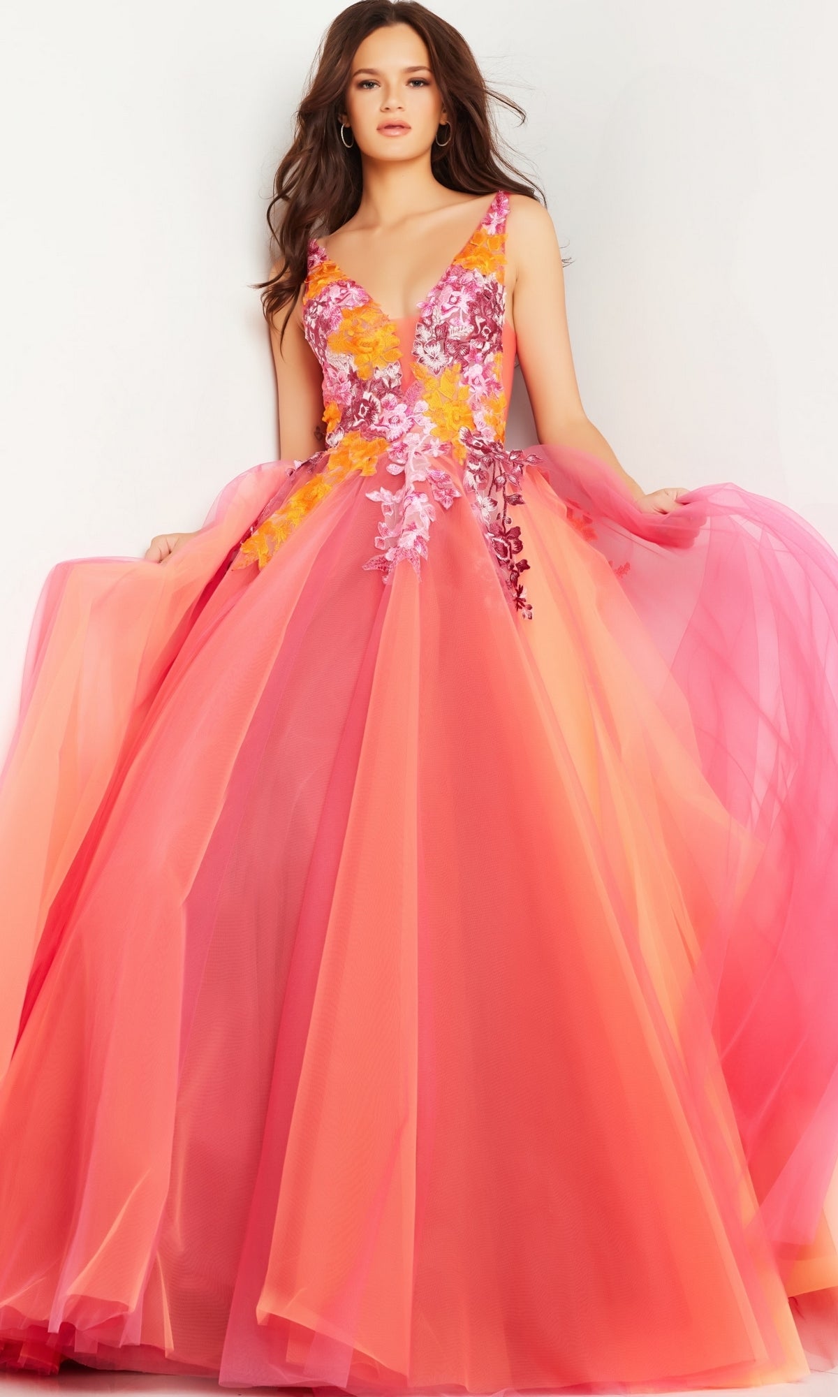 A woman stands against a white background wearing the Jovani V-Neck Long Orange Prom Ball Gown 25800, a sleeveless, floor-length dress with pink and orange floral embroidery and appliqué details on the bodice.