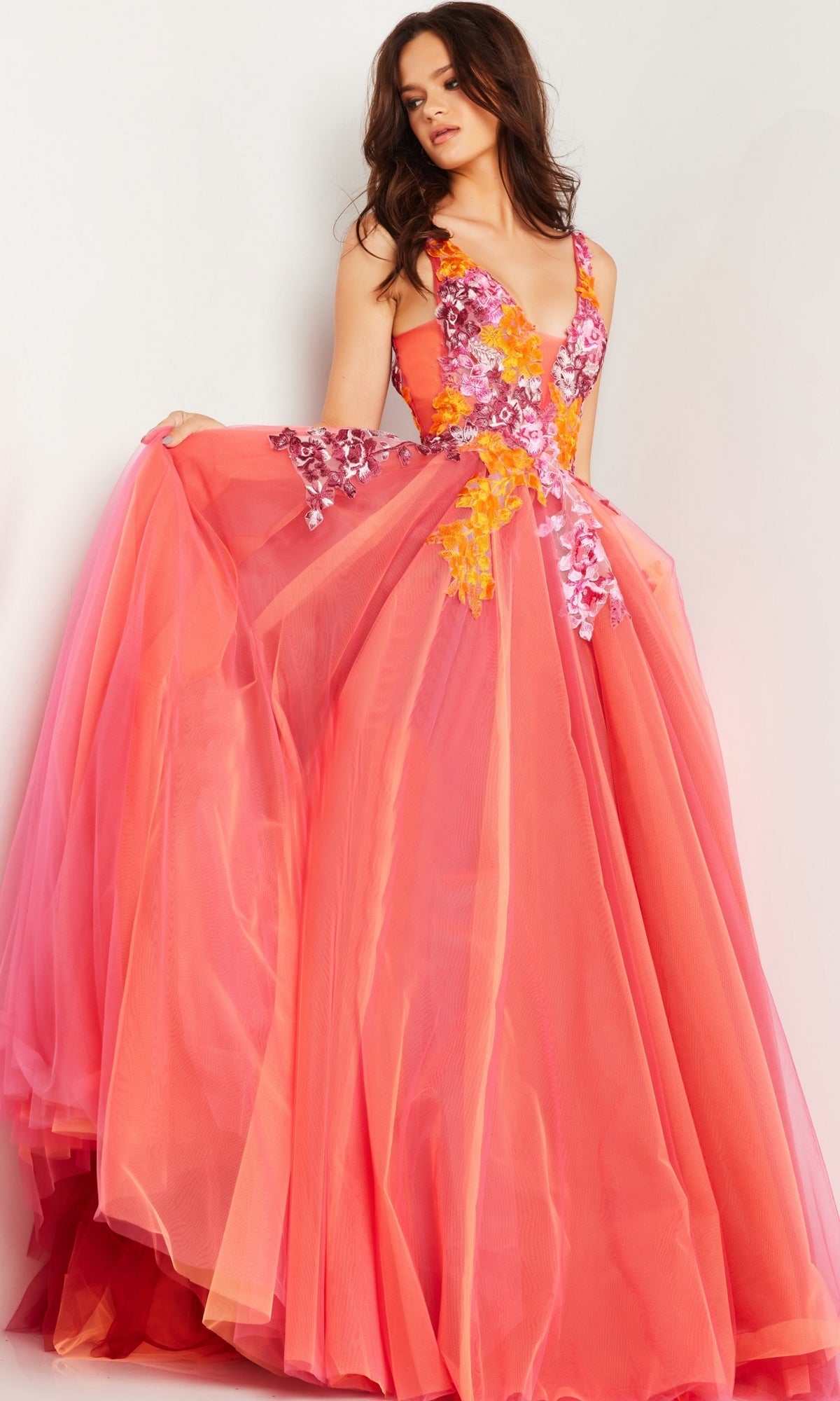 A woman wears the Jovani V-Neck Long Orange Prom Ball Gown 25800, a floor-length, sleeveless A-line dress with floral embroidery on the bodice and a full tulle skirt in pink and orange shades.