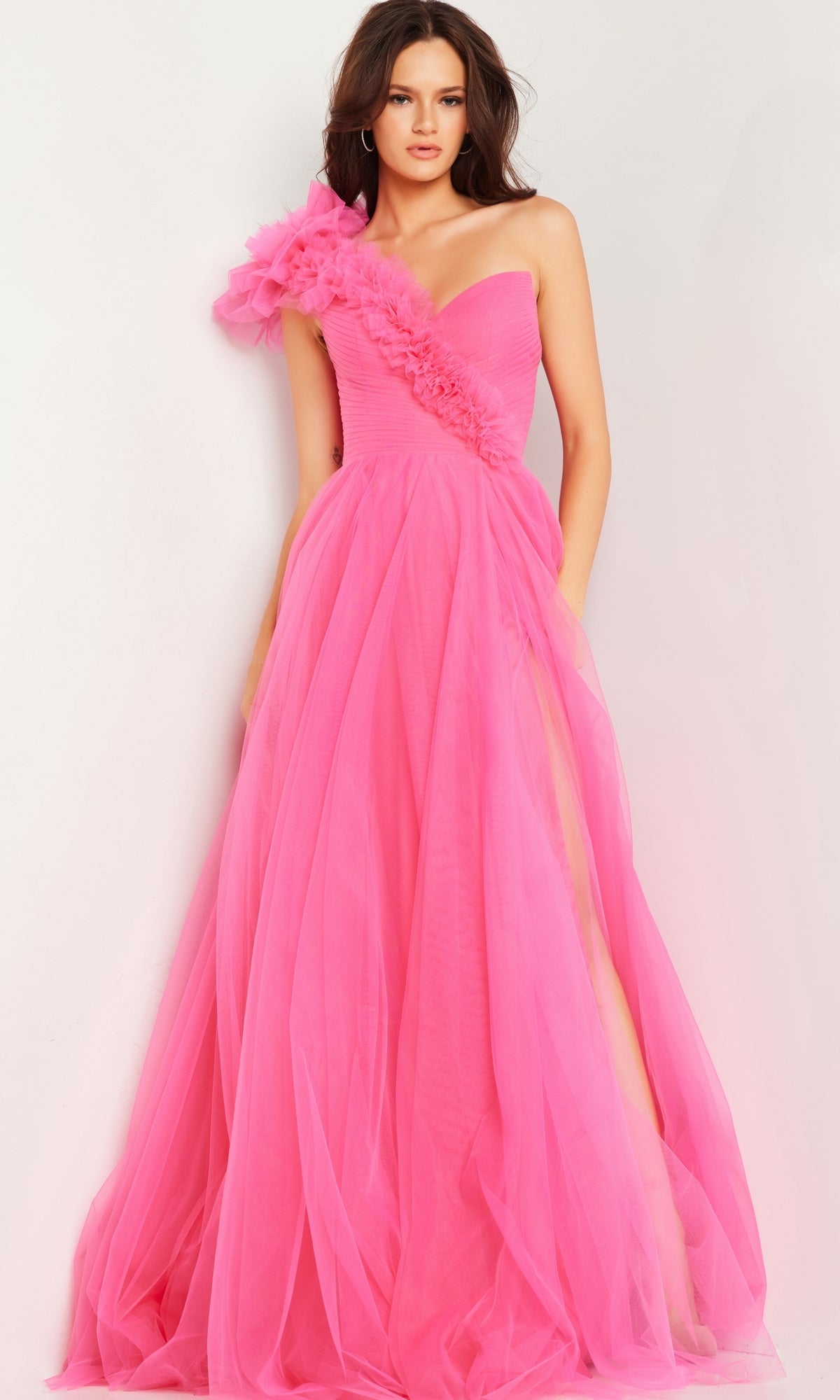 A woman models the Jovani 25919, a bright pink one-shoulder long prom ball gown with tulle and floral appliqué on the shoulder, standing against a plain white background.