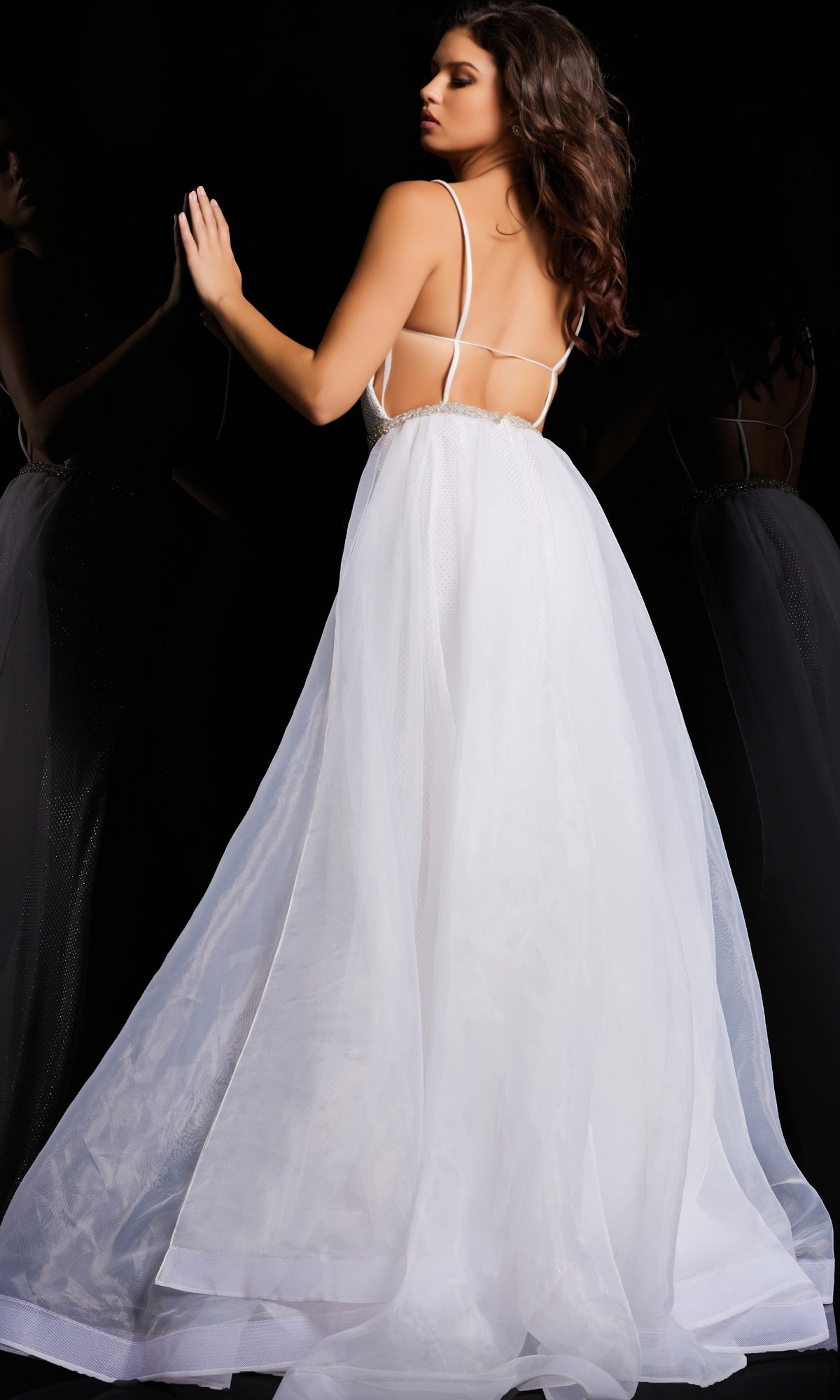 A woman in the Jovani Tulle-Overskirt Beaded-Net Prom Dress 25990, featuring a white backless design with thin straps, stands against a dark background, her hand touching a reflective surface.