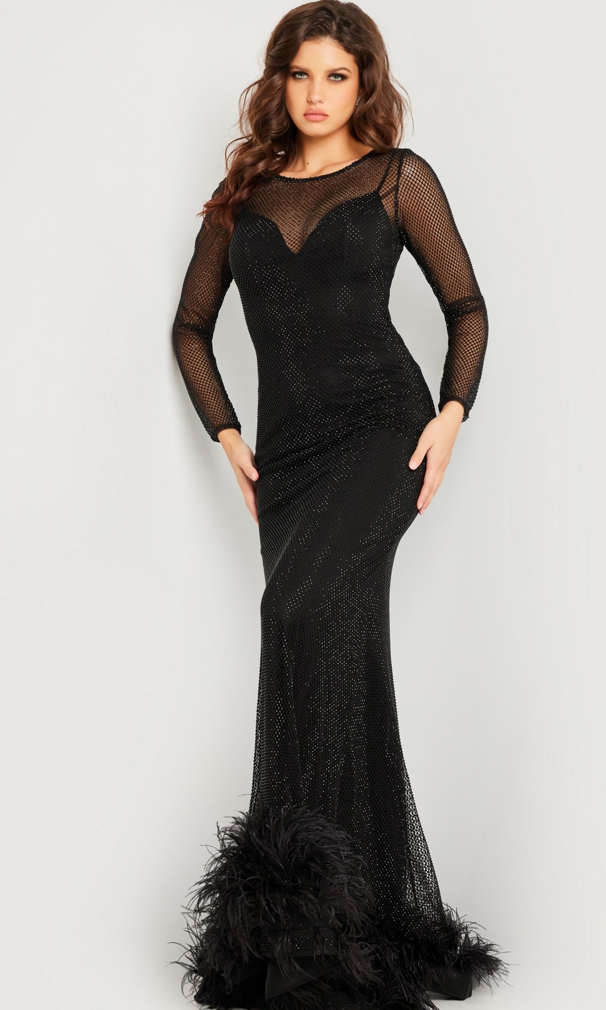 A woman models the Jovani Long Sleeve Long Black Formal Dress 26047, featuring mesh sleeves, a sheer neckline, and feather-accented hem, set against a plain light background.