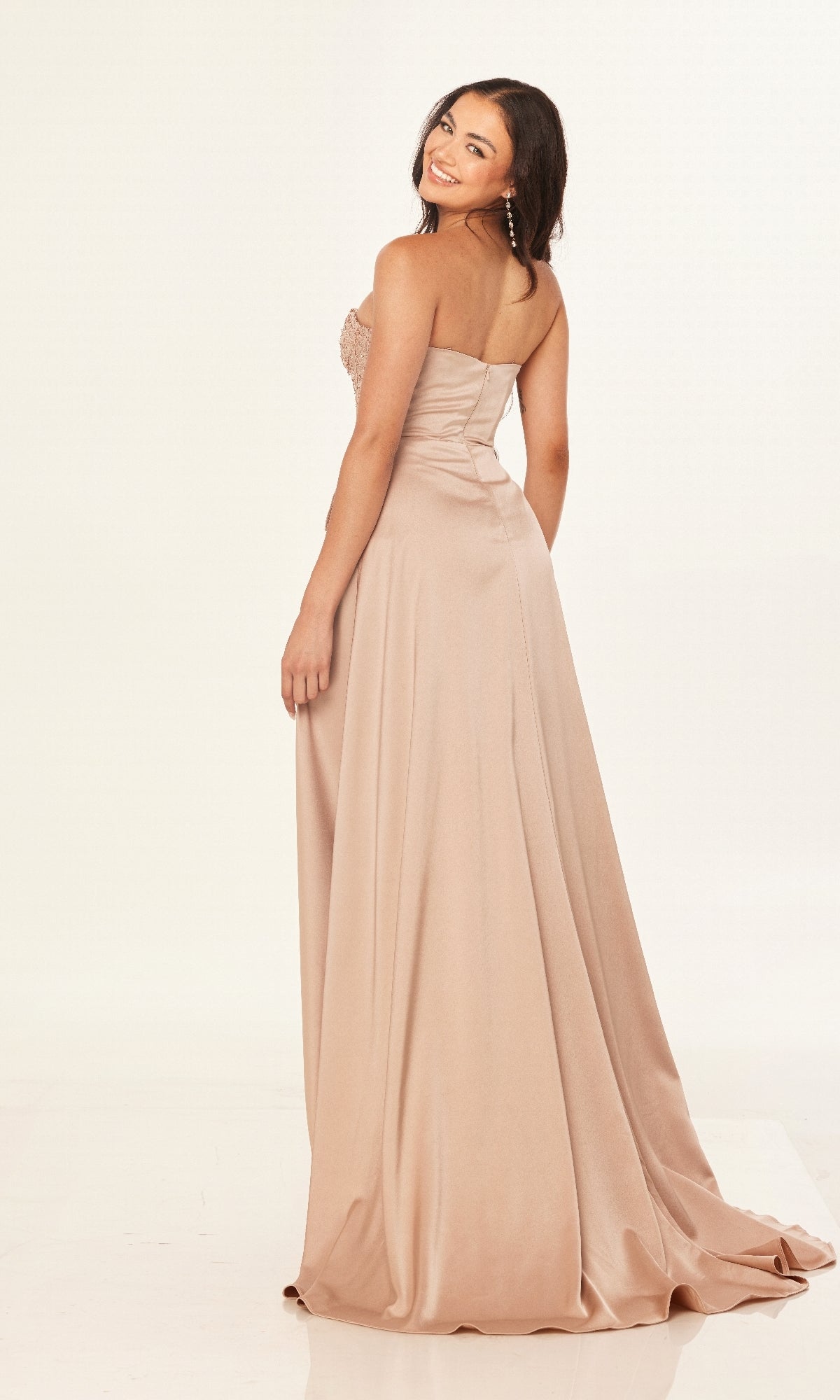 Wearing the Cecilia Couture 2607, a long strapless blush A-line prom dress, a woman stands slightly turned to the side, looking back over her shoulder with a smile.