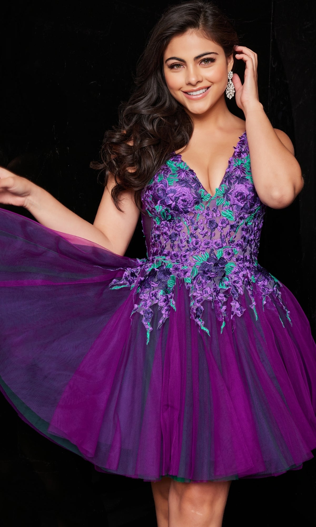 A woman smiles as she holds out the Jovani Short Purple Babydoll Party Dress 26096, featuring floral embroidery and a tulle skirt, against a black background.