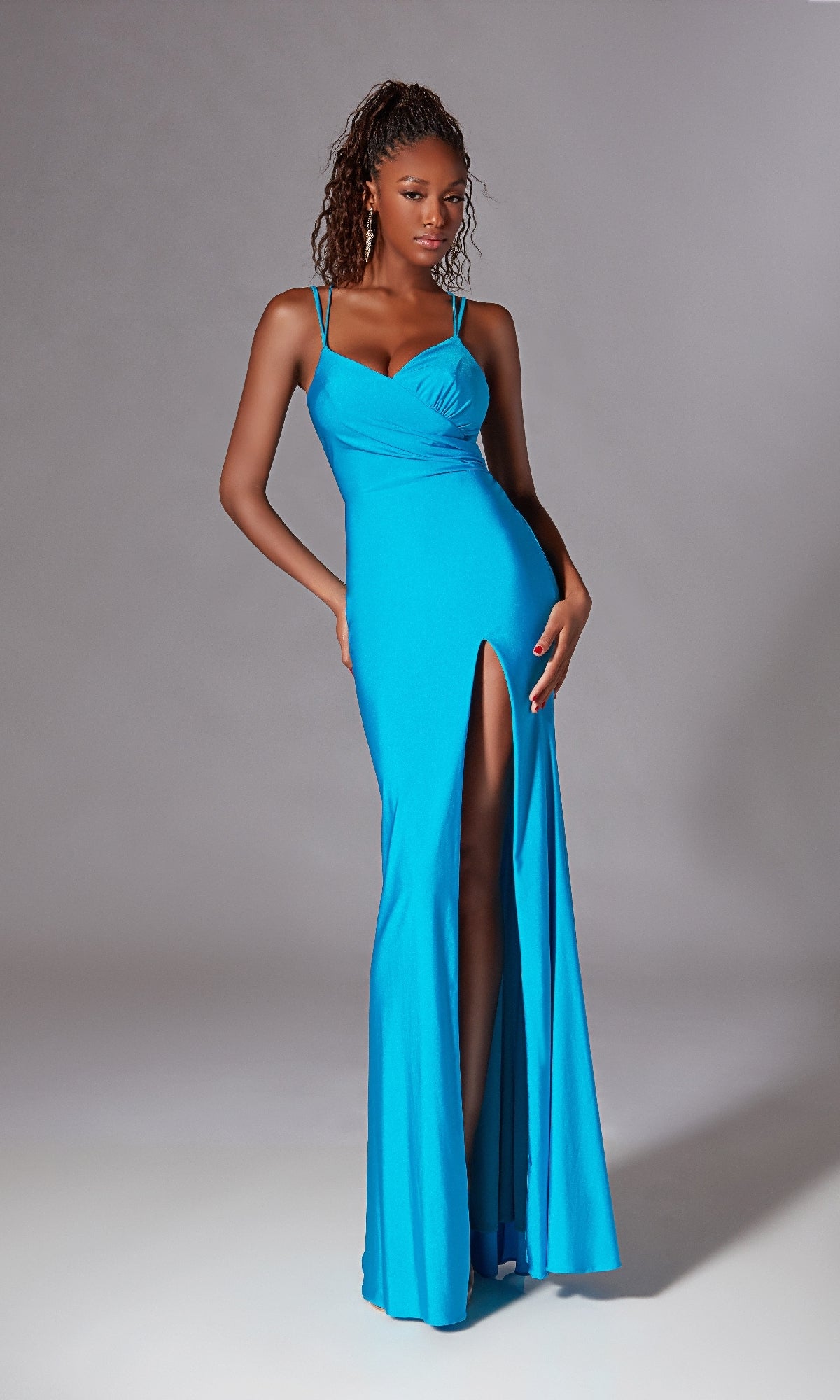 A woman stands against a plain gray background wearing the Aerie Couture 260 Lace-Up Back Long Prom Dress—a bright blue, sleeveless, floor-length gown with spaghetti straps and a high leg slit.