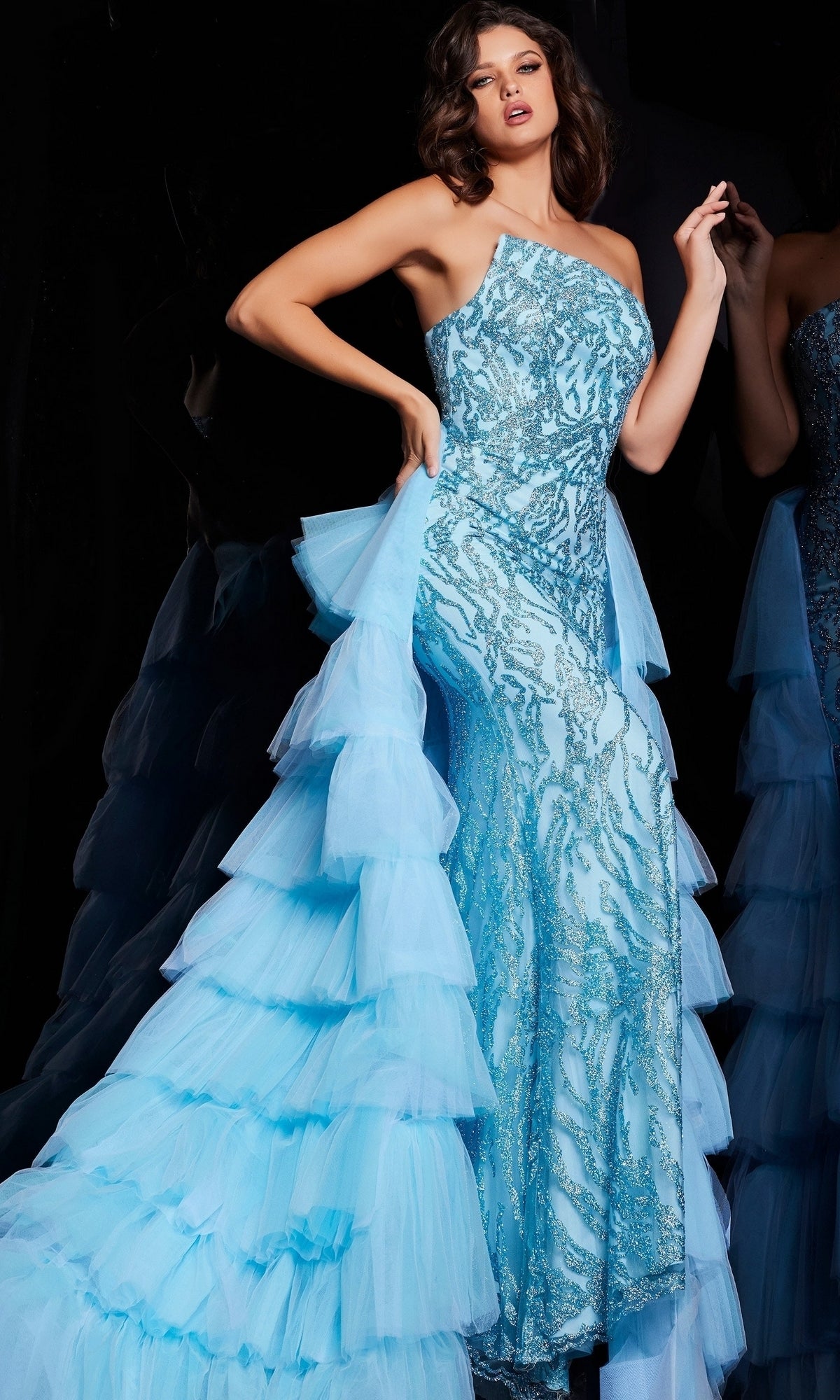A woman poses against a dark background in the Jovani Ruffled Long Glitter-Print Prom Dress 26119, featuring a fitted silhouette, light blue glitter-print fabric, sequins, and a dramatic ruffled tulle train.
