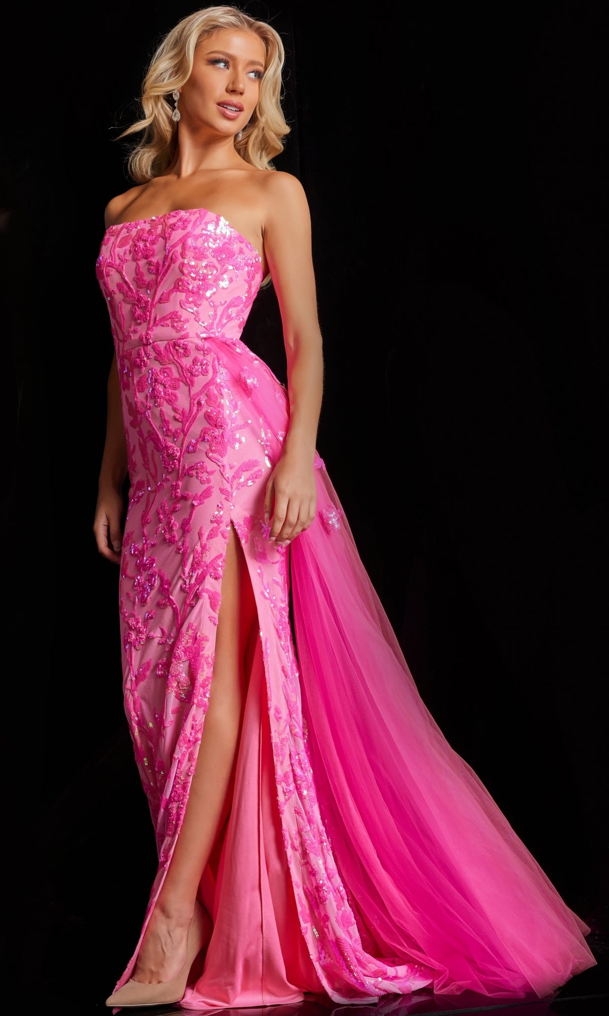 A woman wearing the Jovani Strapless Long Neon Sequin Prom Dress 26134—a floor-length, strapless pink gown with sequin details, a thigh-high slit, and a tulle train—stands against a black background.