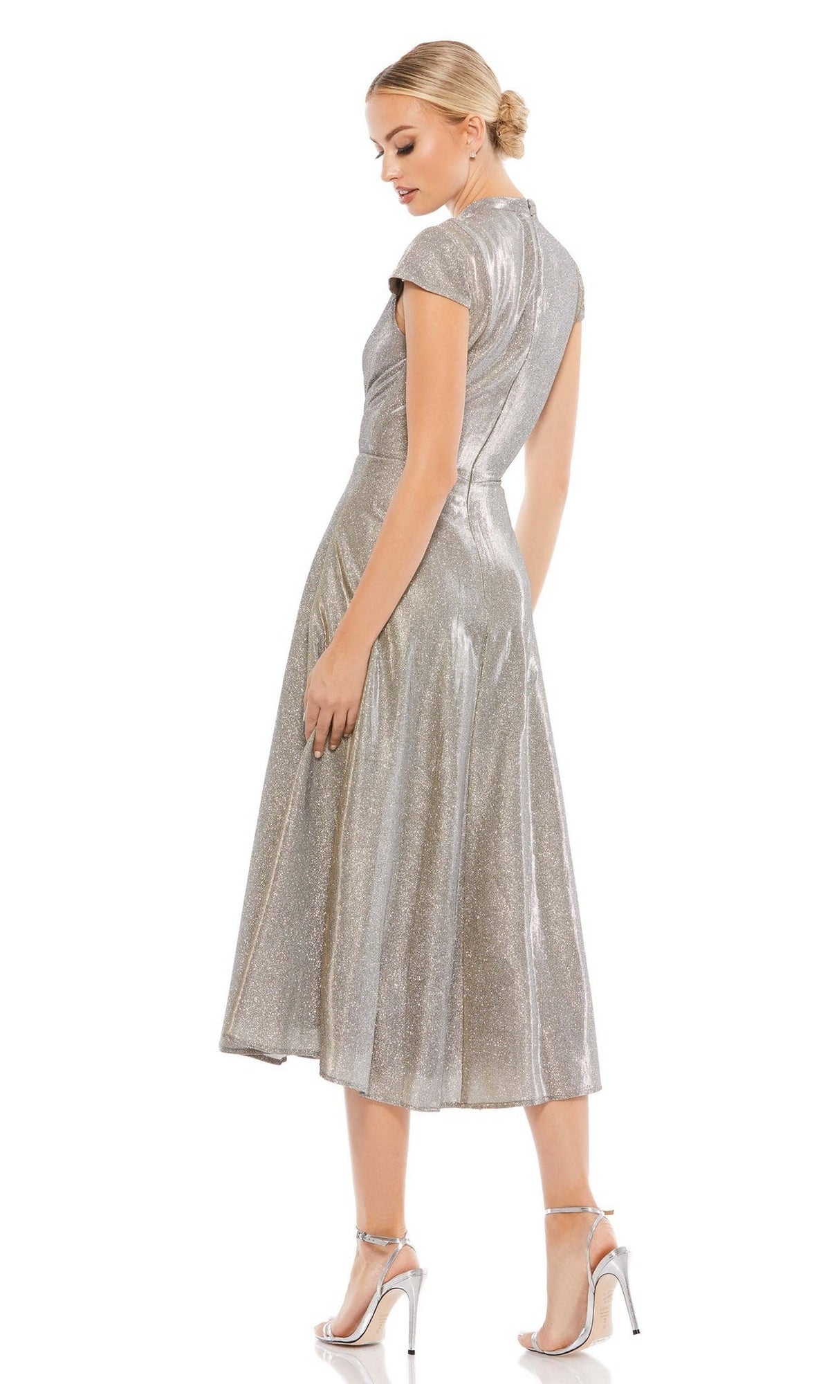 A woman in the Short Wedding Guest Dress 26151 by Mac Duggal, a silver metallic short-sleeved dress, stands turned to the side in silver high-heeled sandals against a white background.