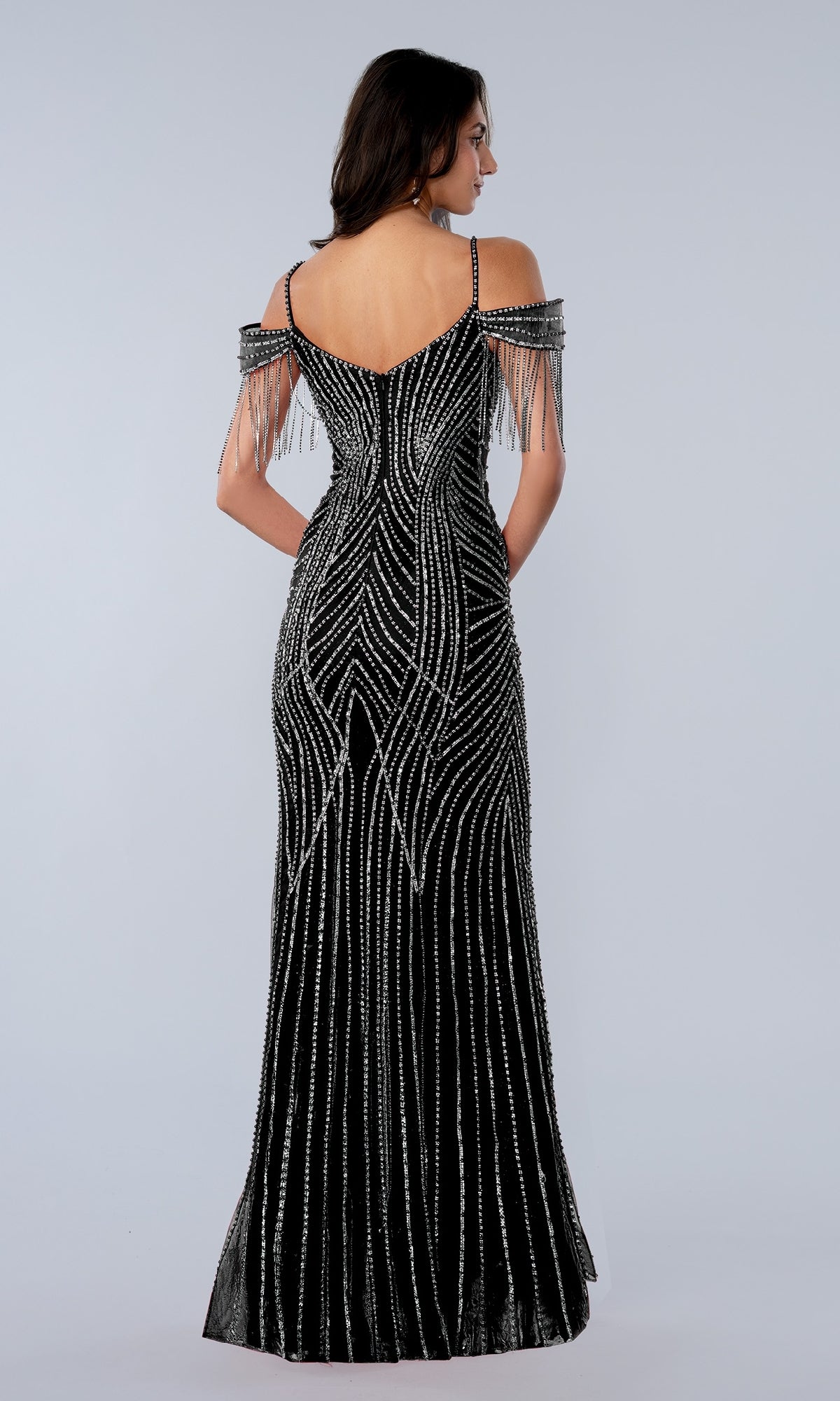 Woman wearing the Long Prom Dress: Stella Couture 26164 in black, featuring off-the-shoulder straps, beaded fringe sleeves, and silver sequin detailing, stands facing away on a plain light background.