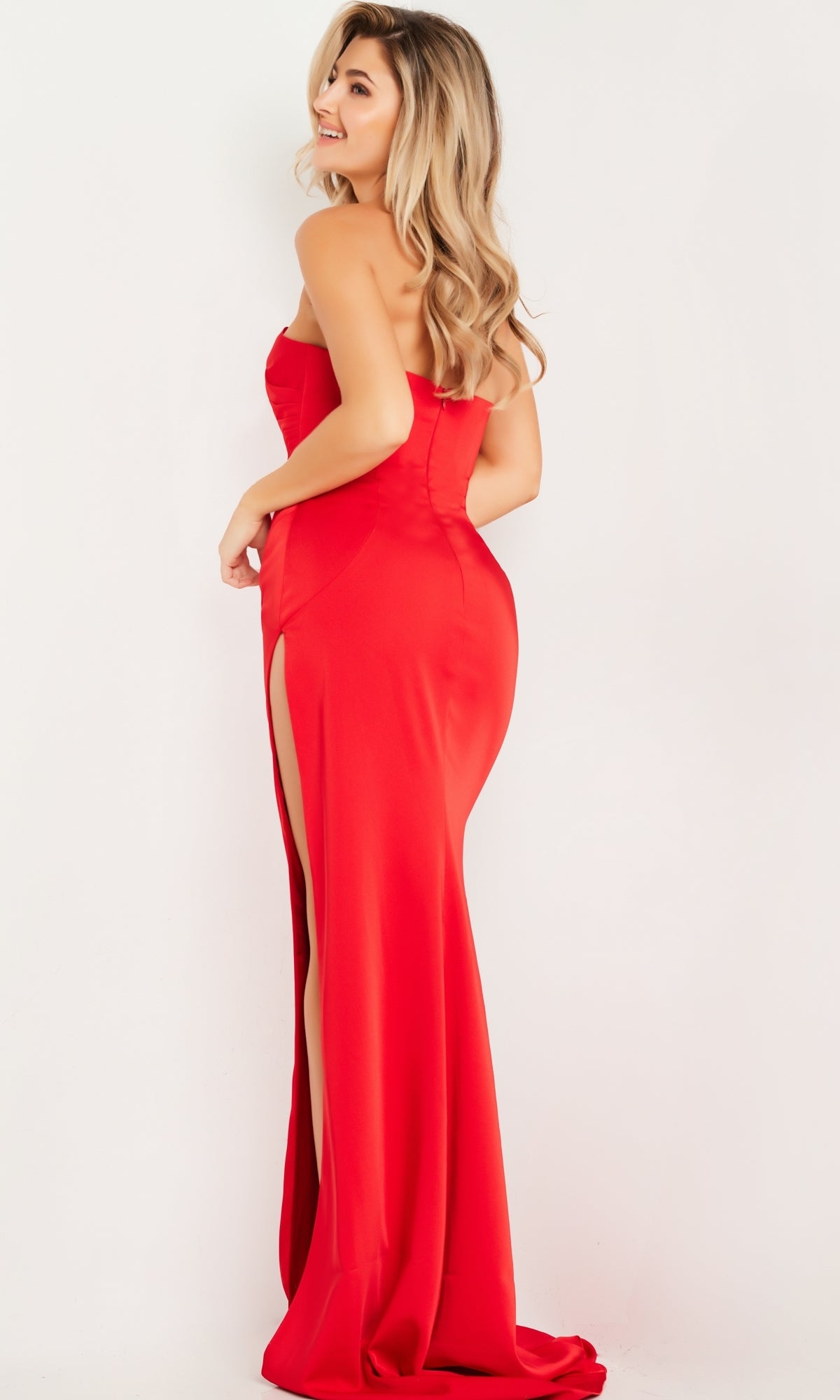 A woman models the Jovani Strapless Long Satin Formal Dress 26166, a red floor-length gown with a side slit, standing sideways and looking over her shoulder against a plain white background.