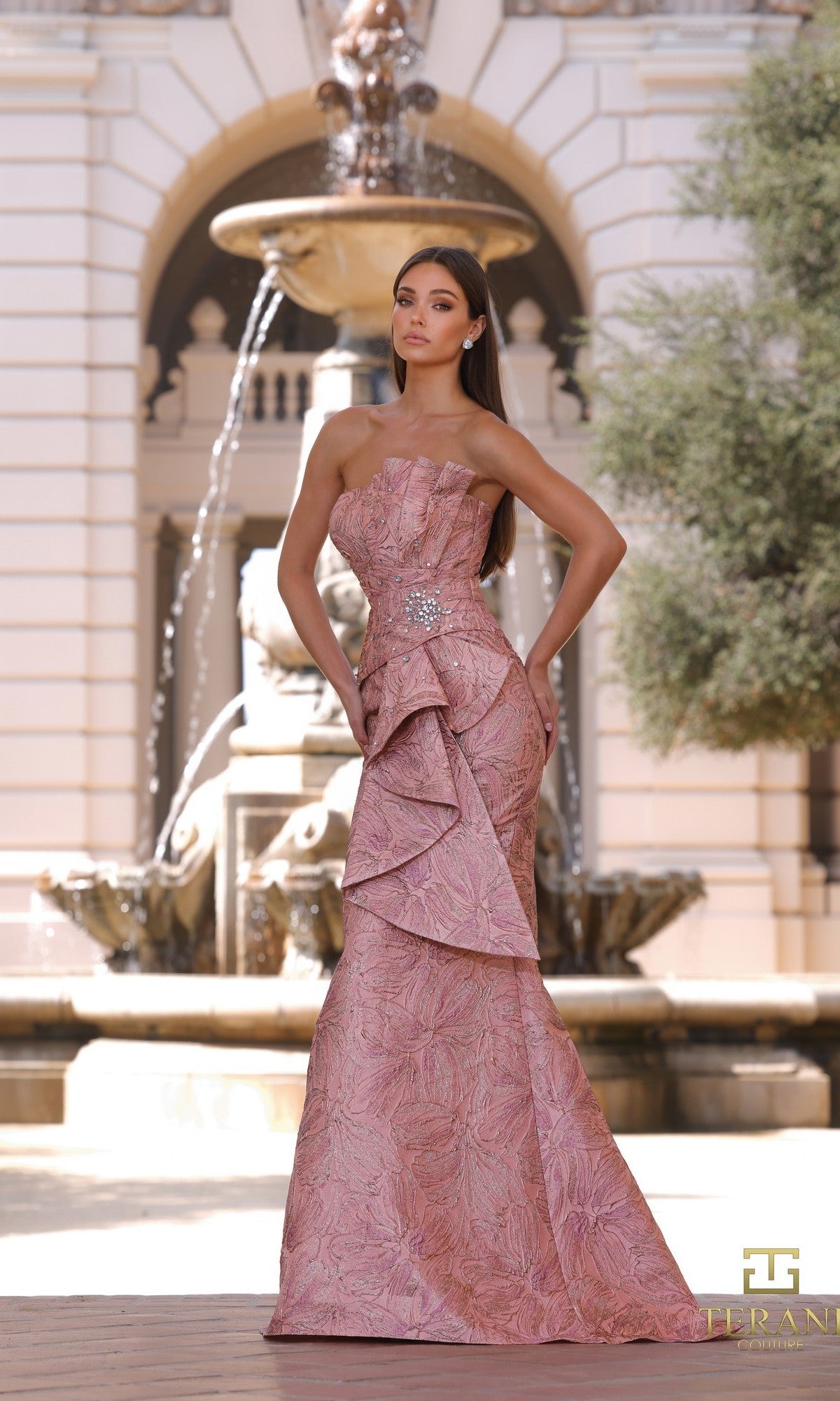 A woman stands outdoors by a fountain, wearing the Terani 261E6269 long formal dress in Blush Rose. This elegant strapless jacquard gown features a structured, asymmetrical design and textured detailing.