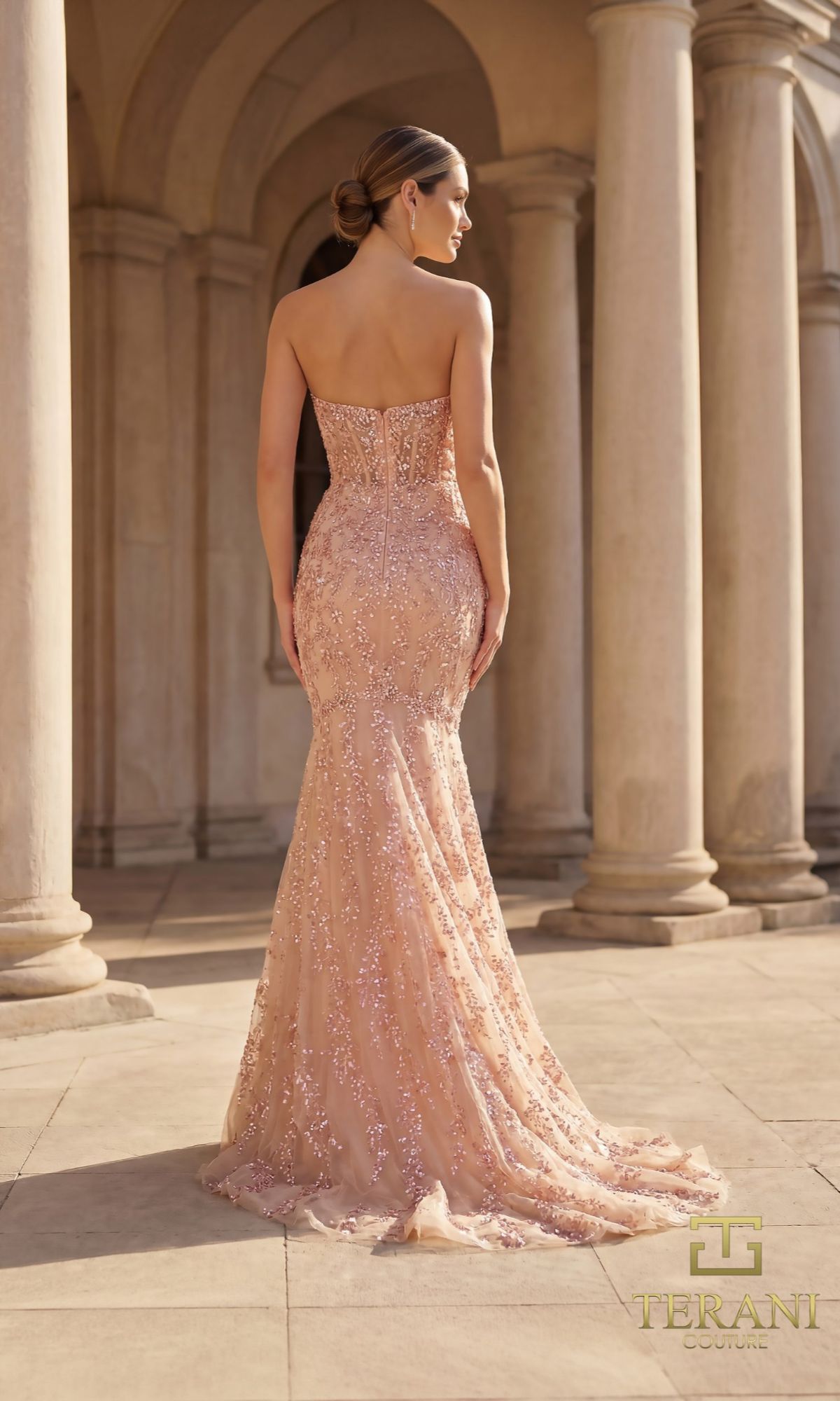 A woman in the Terani 261GL6001 Long Formal Dress stands with her back to the camera, blush pink sequins shimmering on the strapless beaded gown in a columned outdoor setting.