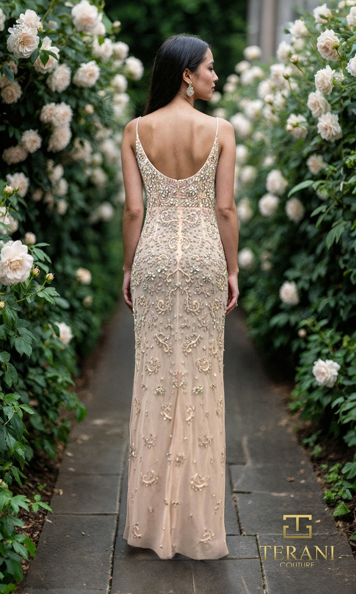 A woman in the Terani 261GL6366 Long Formal Dress stands on a stone path among blooming white roses. The nude, sleeveless beaded tulle sheath gown features a low back and intricate embroidery.