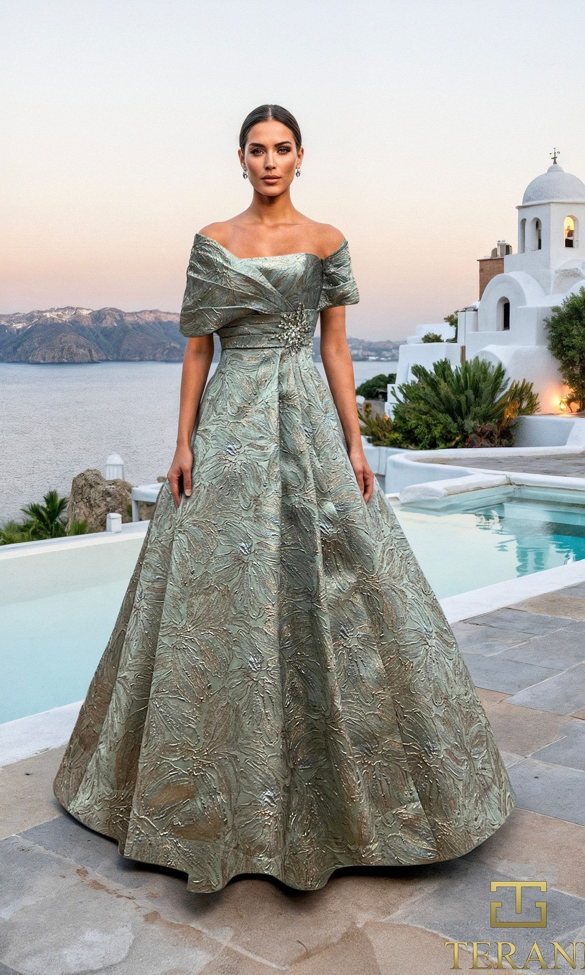 A woman in a seafoam Terani 261M6453 long formal dress stands on a stone patio by a pool, with white buildings and a coastal landscape in the background.