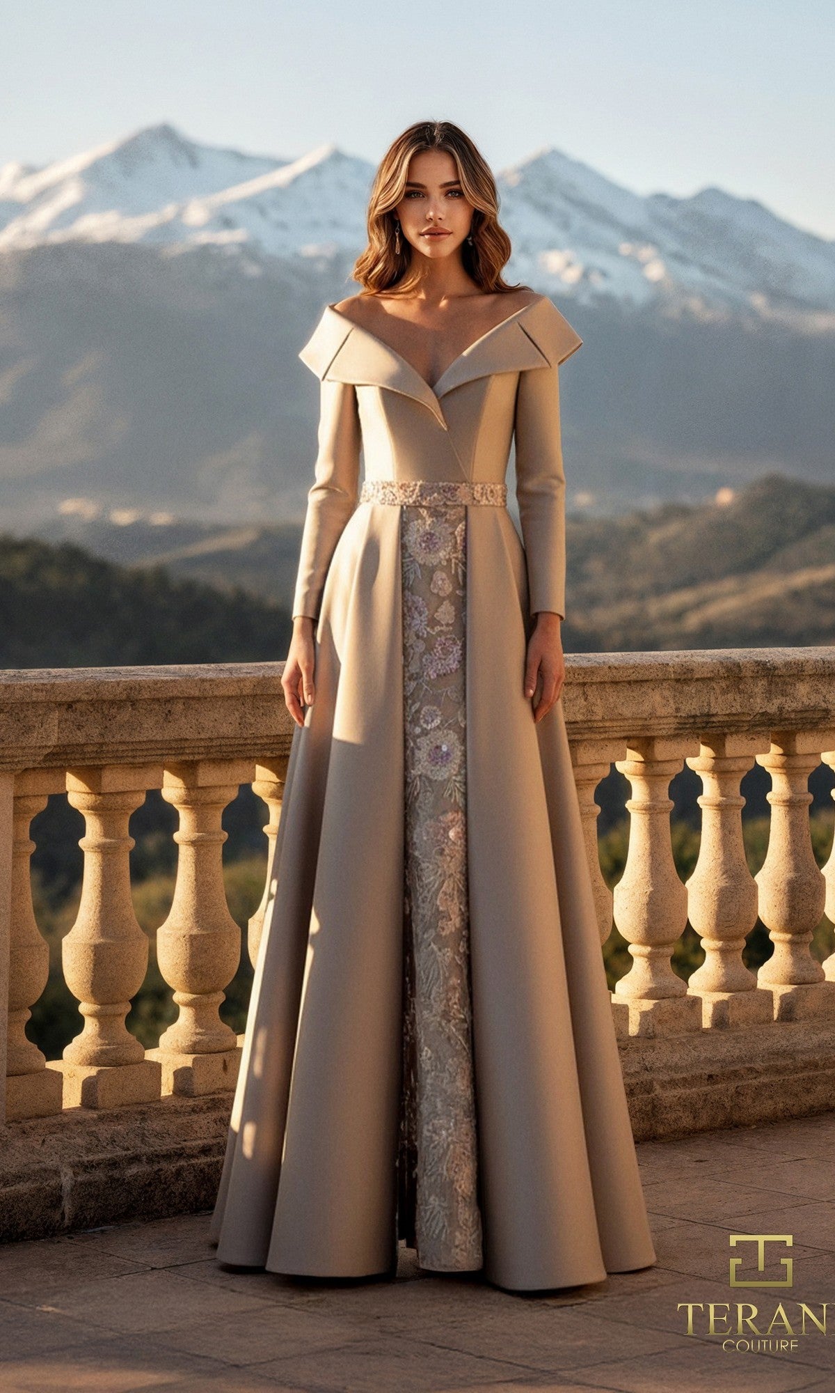 A woman in the taupe Terani 261M6455 long formal dress with lace and Mikado fabric stands on a stone terrace, mountains and clear sky behind her.