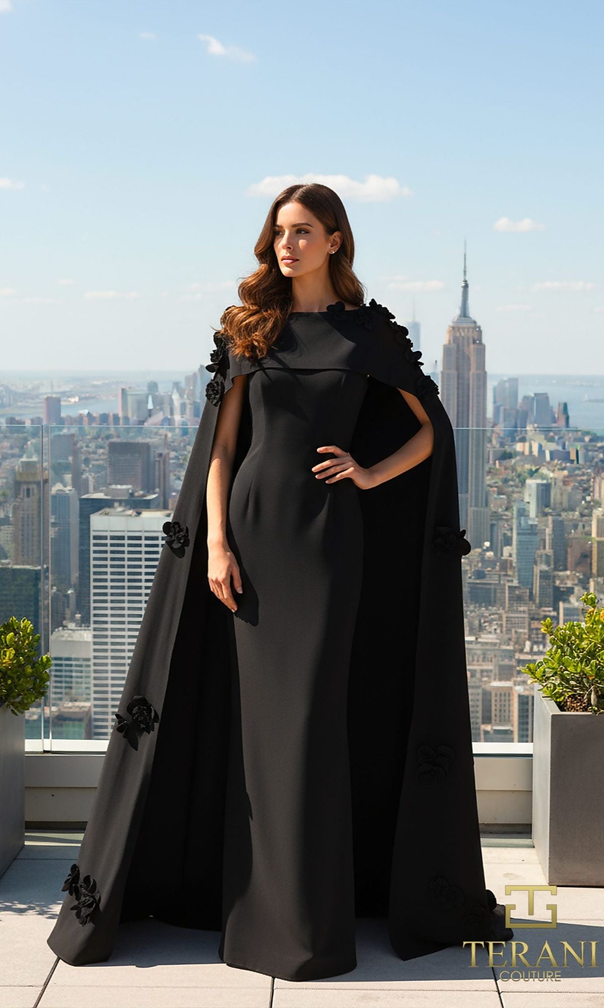 A woman in a long black Terani 261M6687 formal dress with a cape stands on a rooftop, overlooking the city skyline with the Empire State Building in the background.