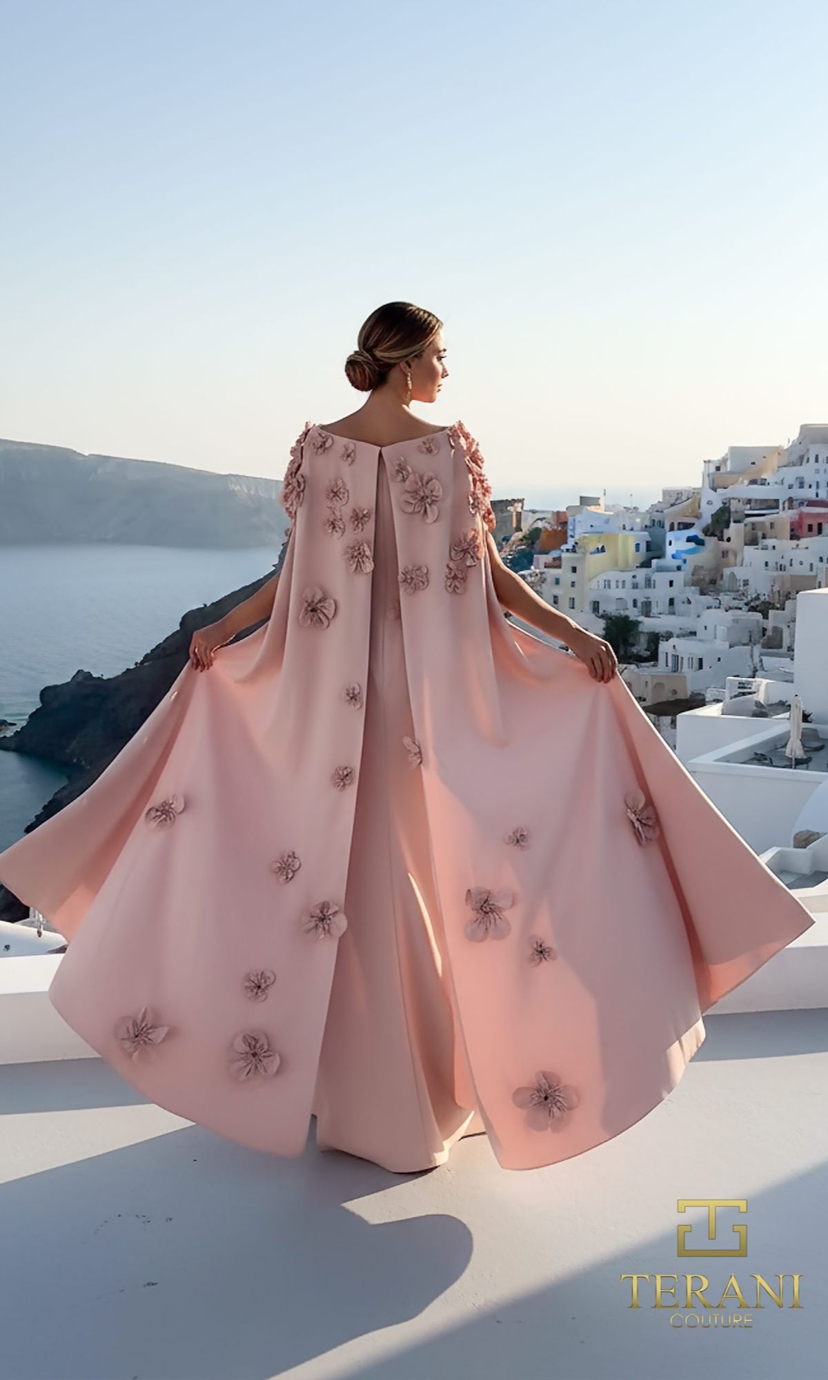 A woman in the Terani 261M6687 long formal dress stands on a terrace, facing away from the camera, overlooking white buildings and a coastline.
