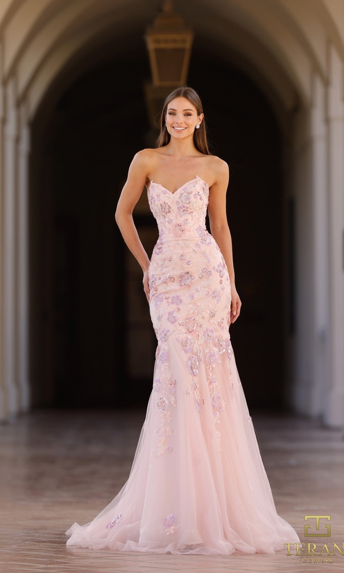 A woman stands and smiles in a covered outdoor walkway wearing the Long Formal Dress: Terani 261P6193, a strapless tulle gown with floral embellishments and a slight mermaid silhouette.