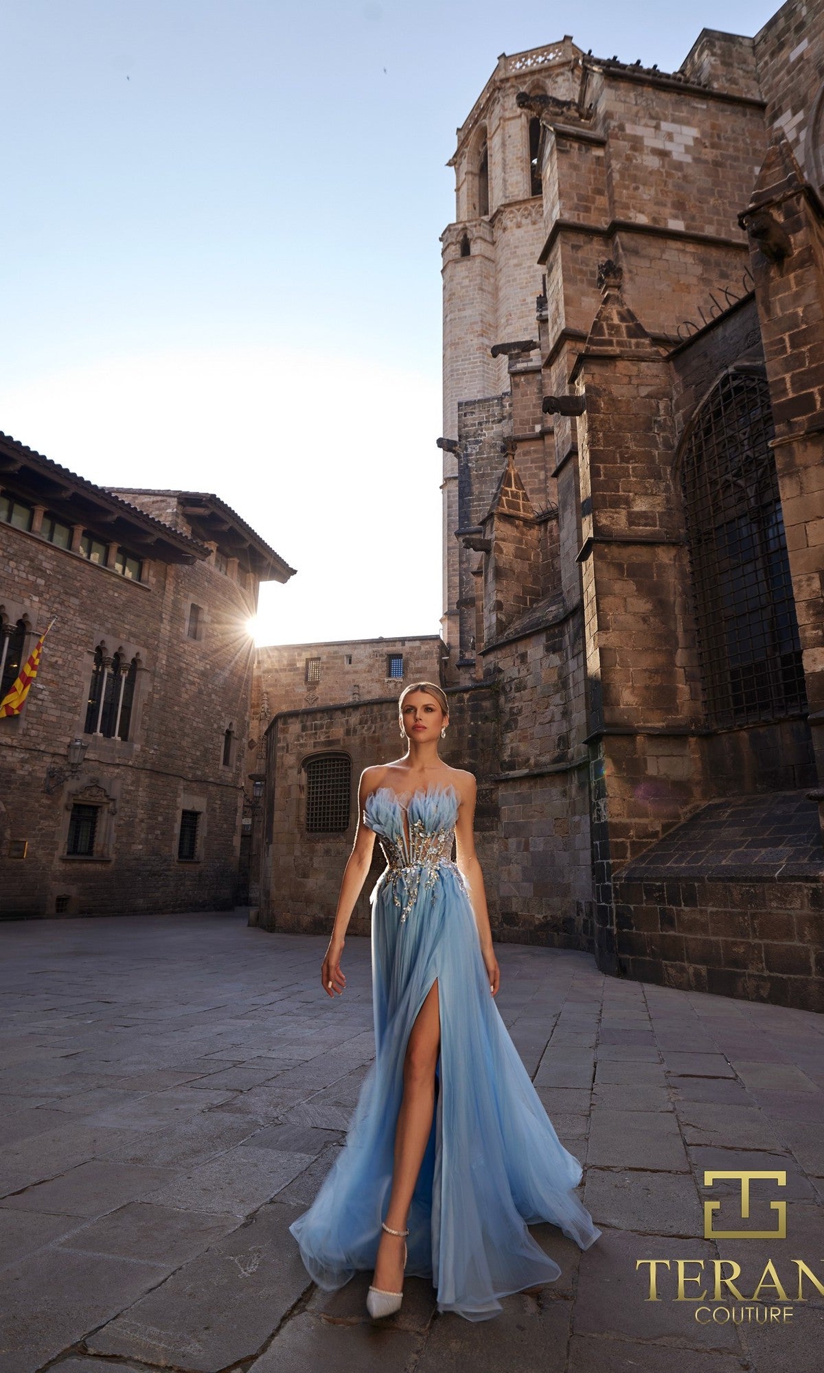 A woman in a flowing blue Terani 261P6631 long formal dress with a high slit walks on a stone courtyard near historic buildings, with the Terani Couture logo in the corner.