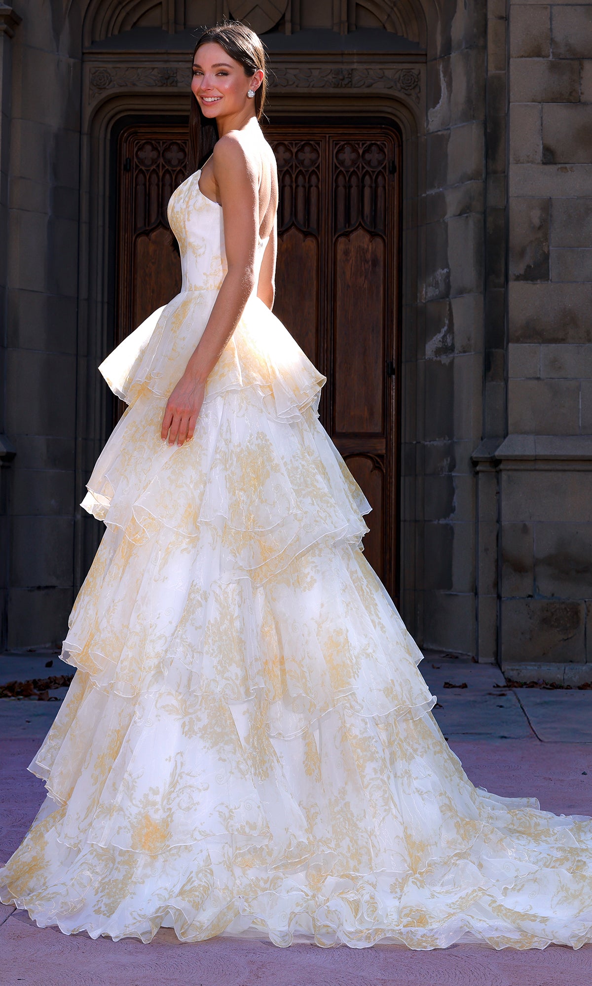 A woman stands outside smiling over her shoulder in the Terani 261P6673 long formal dress, a strapless ball gown featuring tiered layers, a subtle yellow floral pattern, and an A-line chiffon skirt.