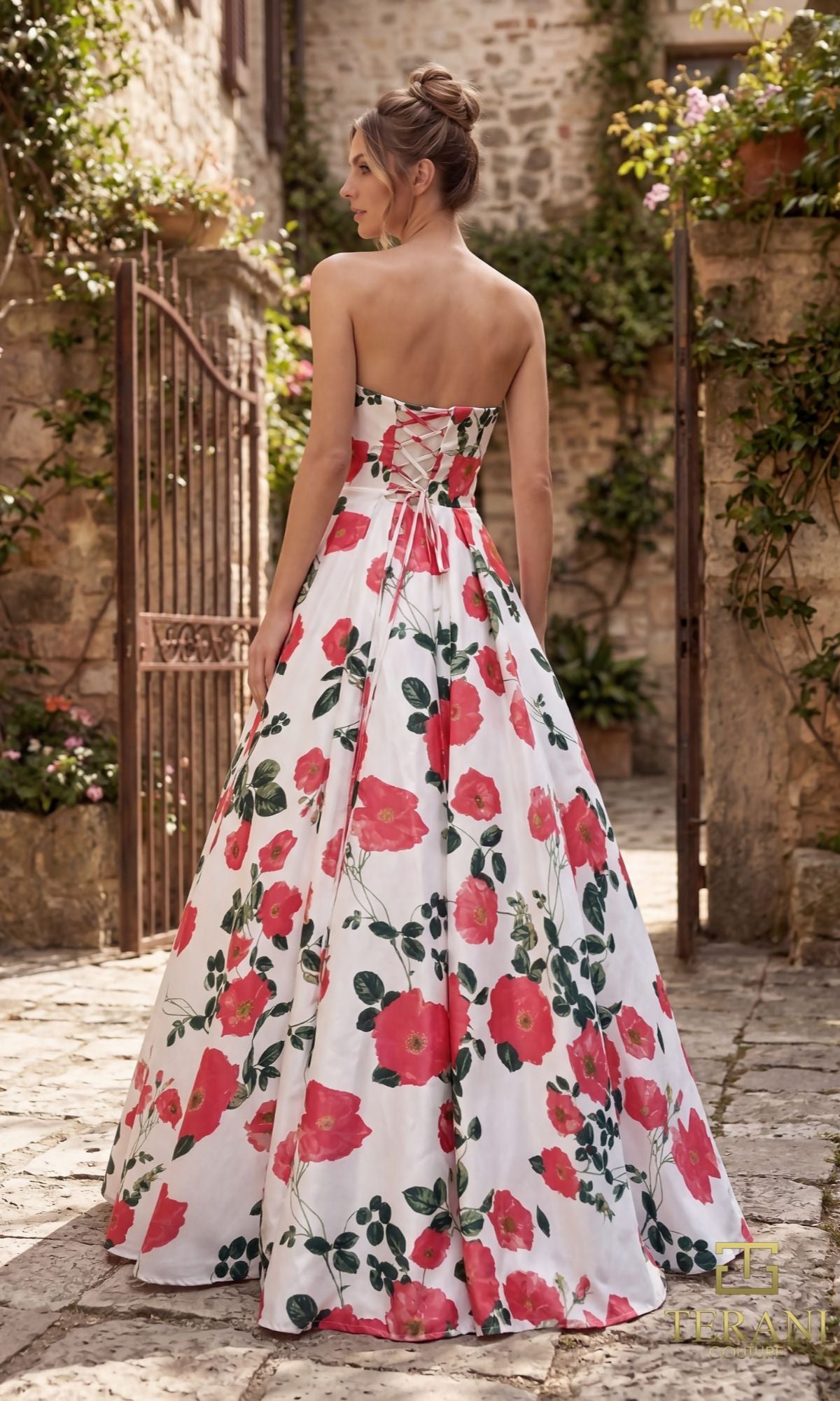 A woman stands outdoors with her back to the camera, wearing the strapless Terani 261P6705 long formal dress. Stone walls, plants, and an open gate are visible in the background.