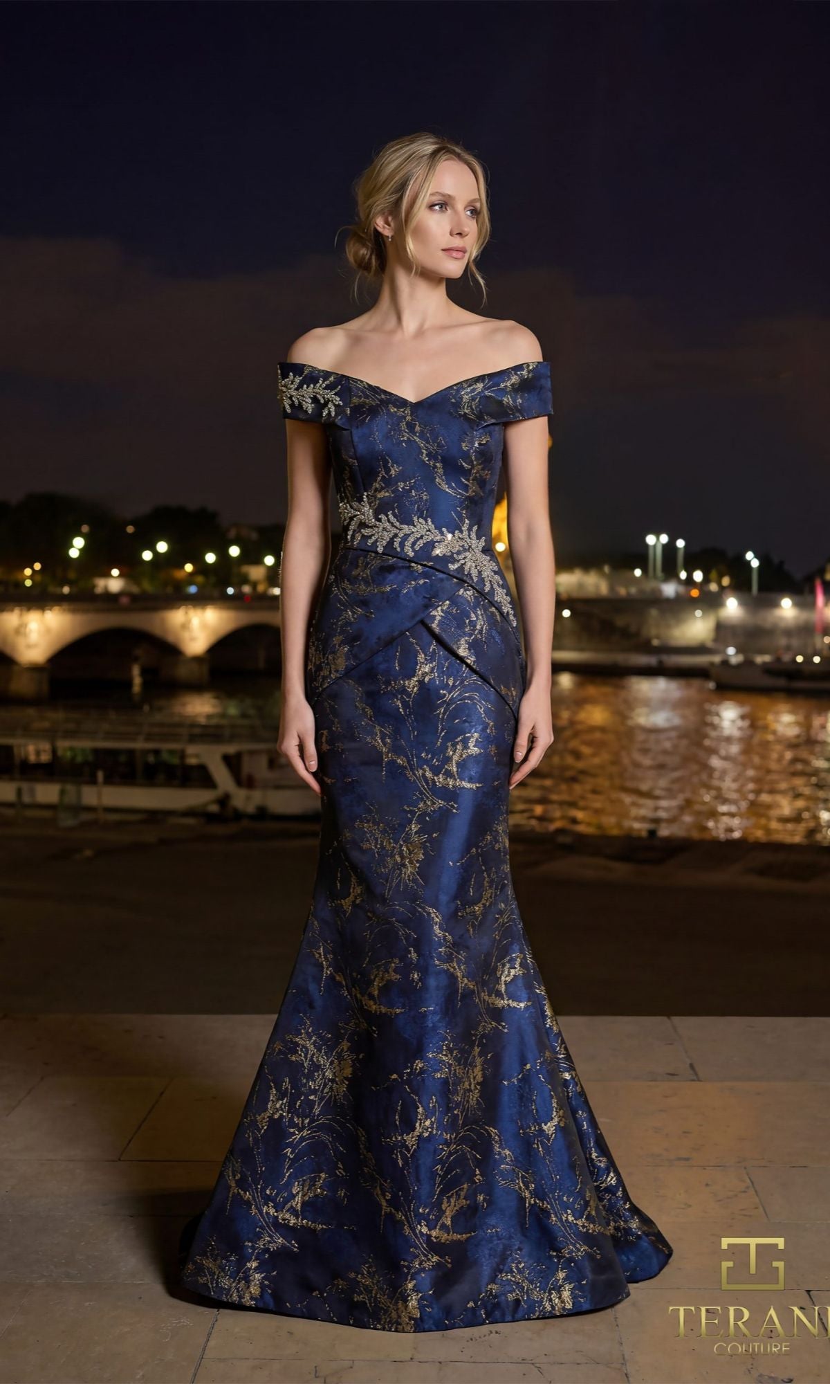 A woman stands outdoors at night in the Terani 261E6252 long formal dress, a floor-length midnight gold gown. City lights and a bridge over water provide a stunning backdrop.