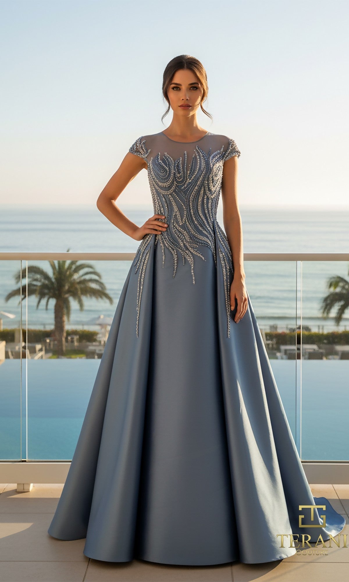 A woman in a slate Long Formal Dress: Terani 261E6254, crafted from stretch Mikado with silver embellishments, stands on a balcony overlooking the sea, with palm trees and a pool in the background.