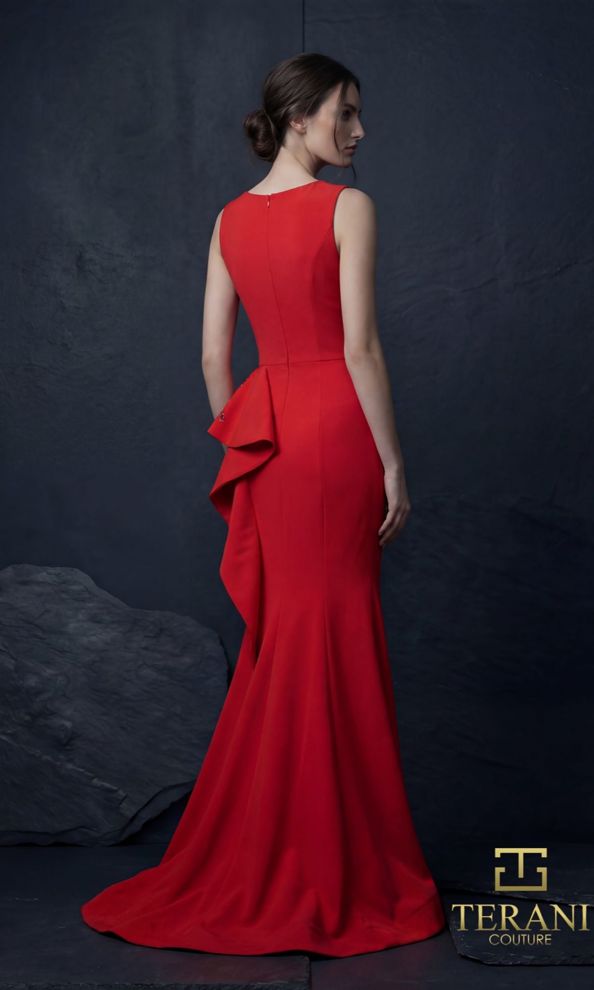 A woman wears the Long Formal Dress: Terani 261E6750?a fitted, floor-length red gown with ruffled back detail and a mermaid silhouette?standing against a dark textured background.