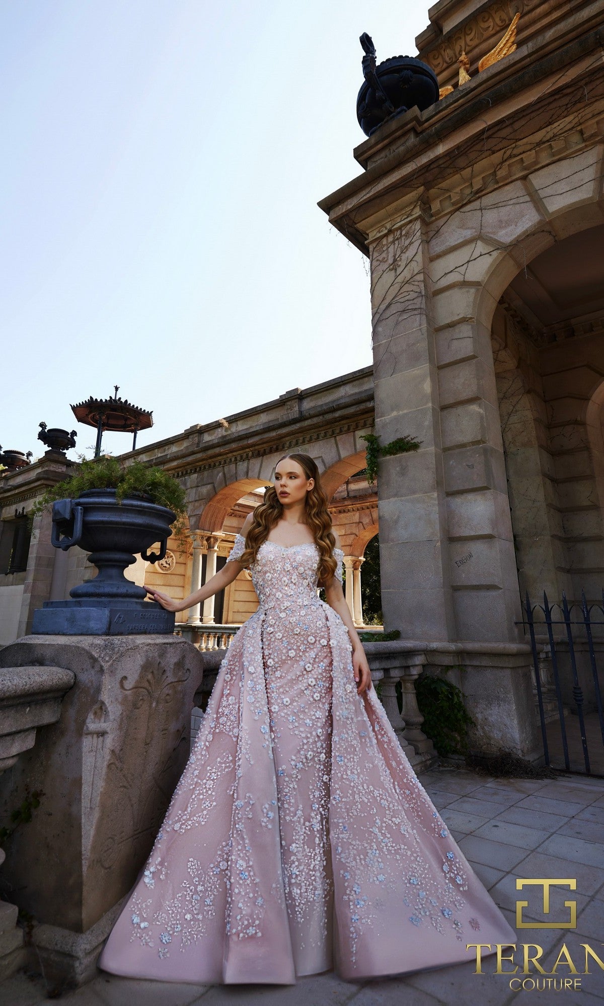 A woman in a pink blue Terani 261GL6659 long formal dress with delicate floral embellishments stands outdoors by a classical stone building, her hand resting on a planter.