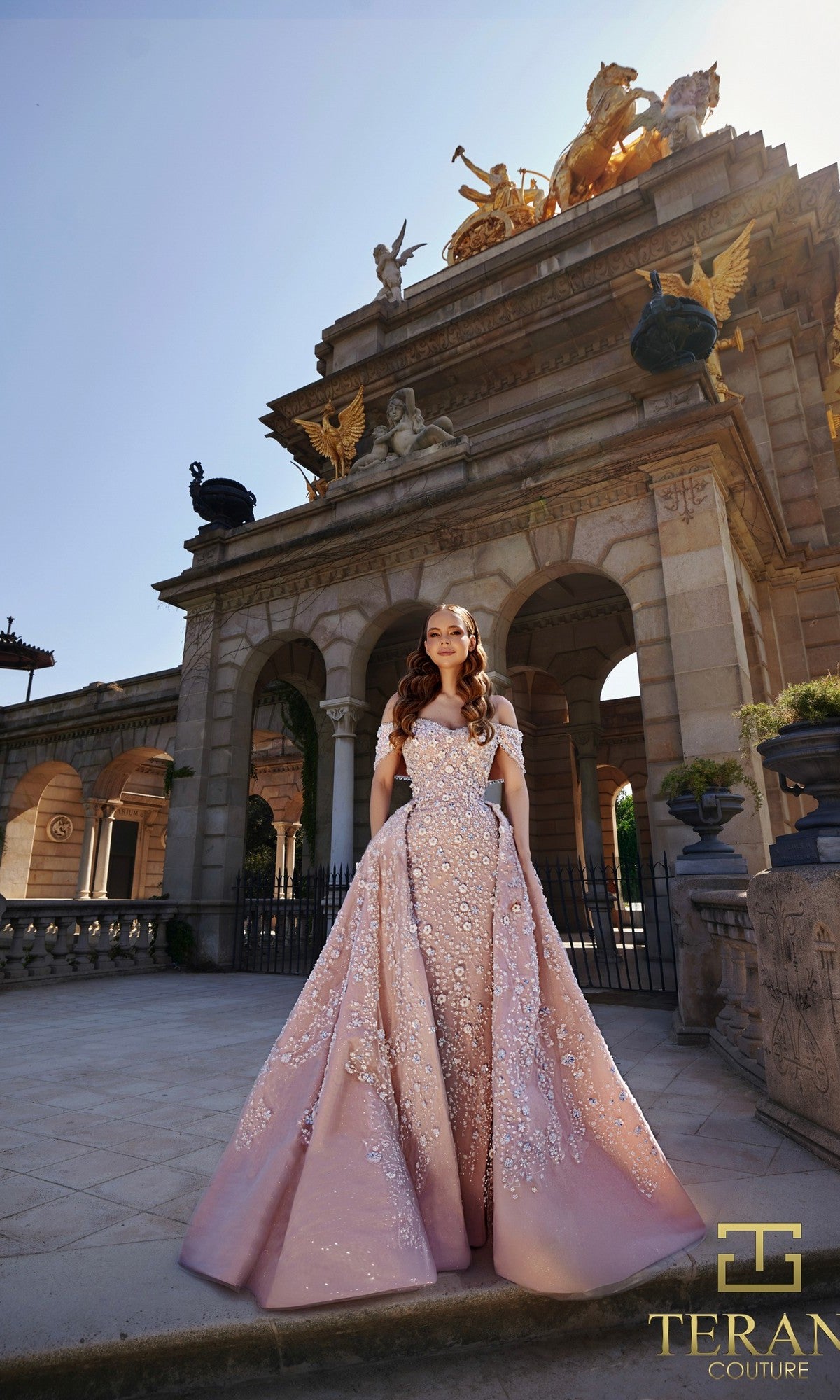 A woman in a pink embroidered tulle Terani 261GL6659 formal dress poses gracefully before an ornate stone monument with statues and arches. The Terani Couture logo appears in the corner.