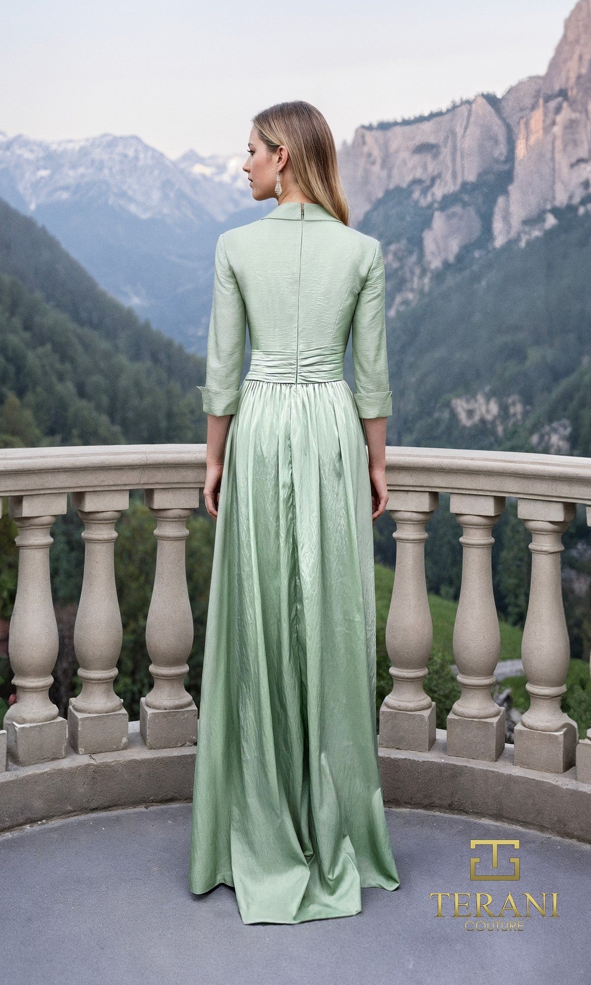 A woman in the Terani 261M6418 long formal dress, made of matte taffeta in light green, stands on a stone balcony facing away and overlooking mountains. The Terani Couture logo appears at the bottom right.