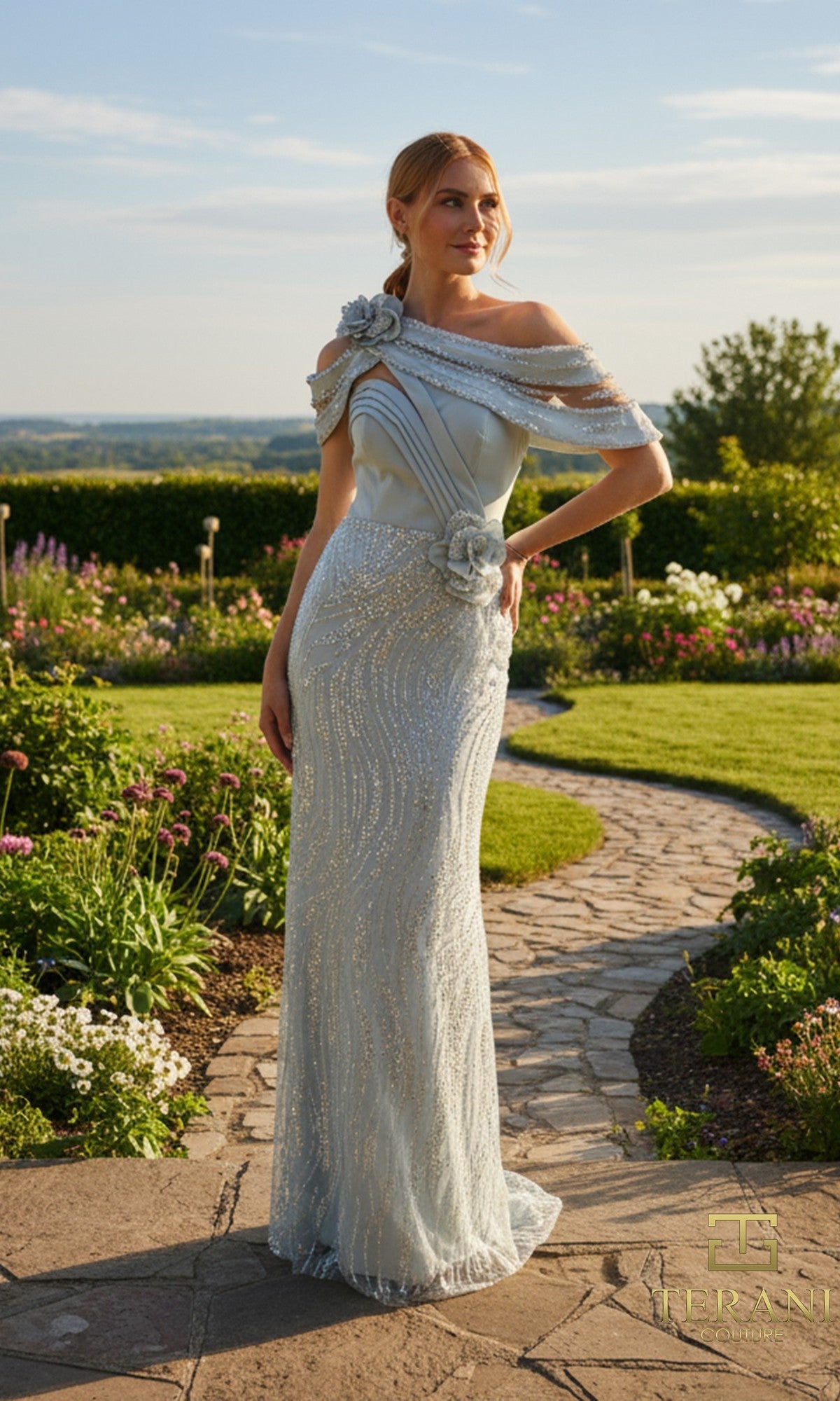 A woman in a sage, off-shoulder Terani 261M6427 gown with floral embellishments and a beaded tulle sheath skirt stands on a stone path in a landscaped garden.