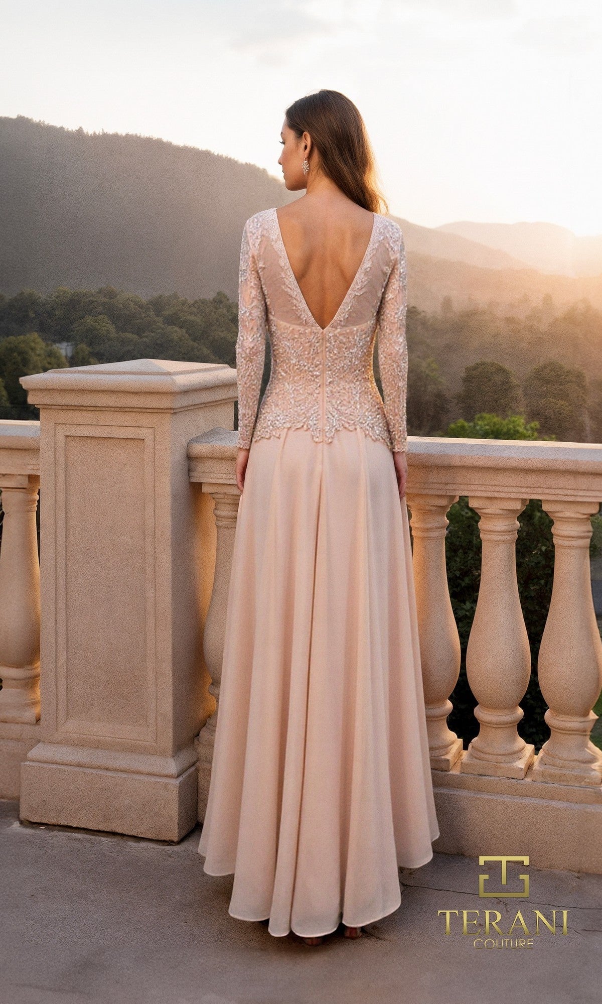 A woman in the Long Formal Dress: Terani 261M6439, featuring lace sleeves, a deep V-back, and an elegant a-line chiffon skirt, stands on a stone balcony overlooking a scenic sunset landscape.