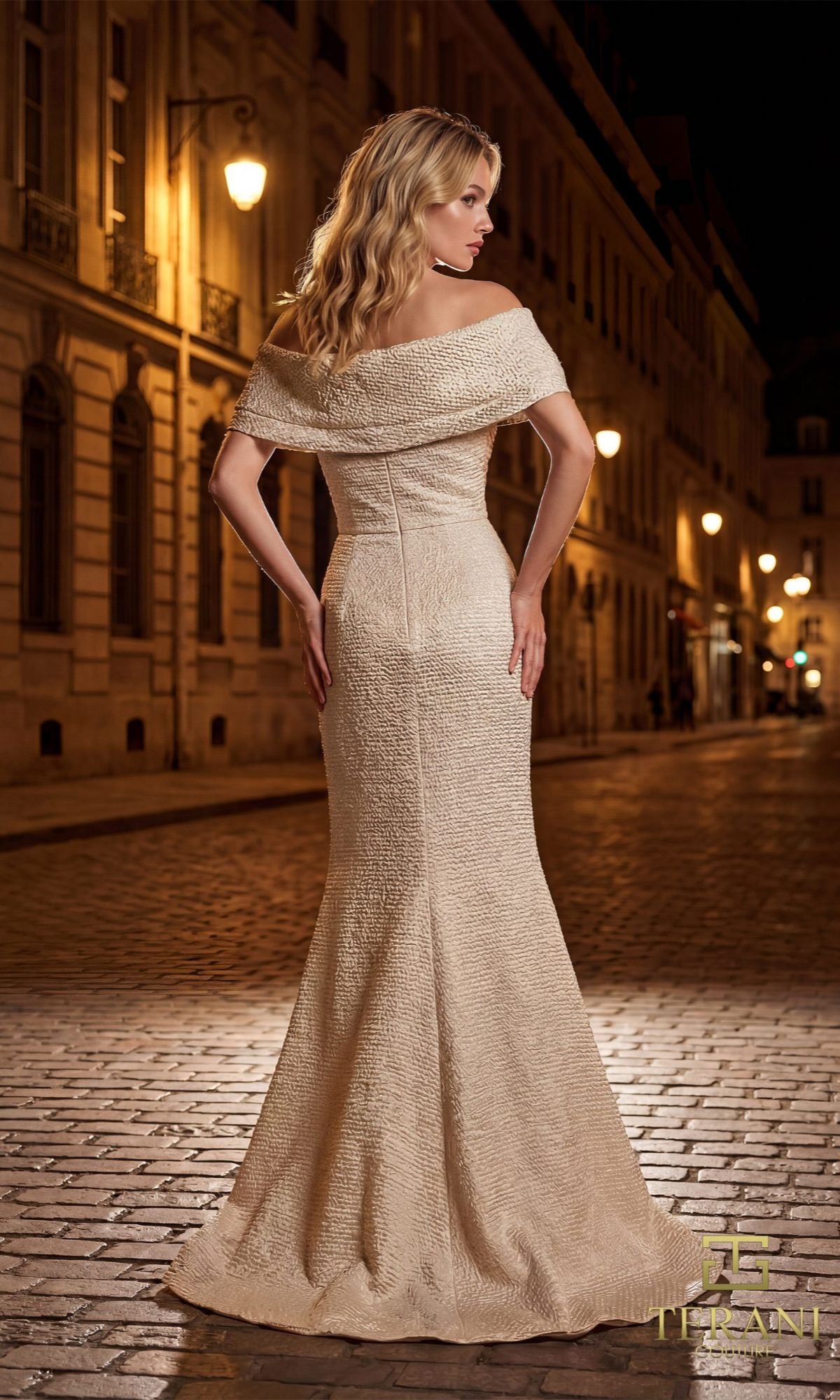 A woman in a cream, off-shoulder Terani 261M6449 evening gown stands on a cobblestone street at night, her look echoing the elegance of a metallic jacquard mother-of-the-bride dress as she faces away from the camera.