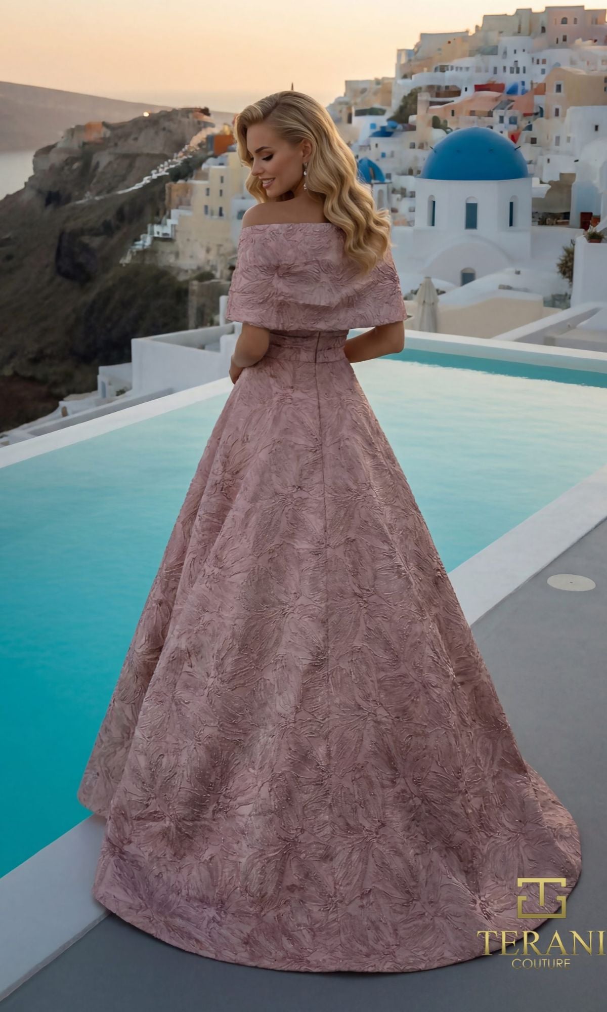 A woman in a blush rose Long Formal Dress (Terani 261M6453) stands by a pool, with the white buildings and blue domes of Santorini in the background at sunset.