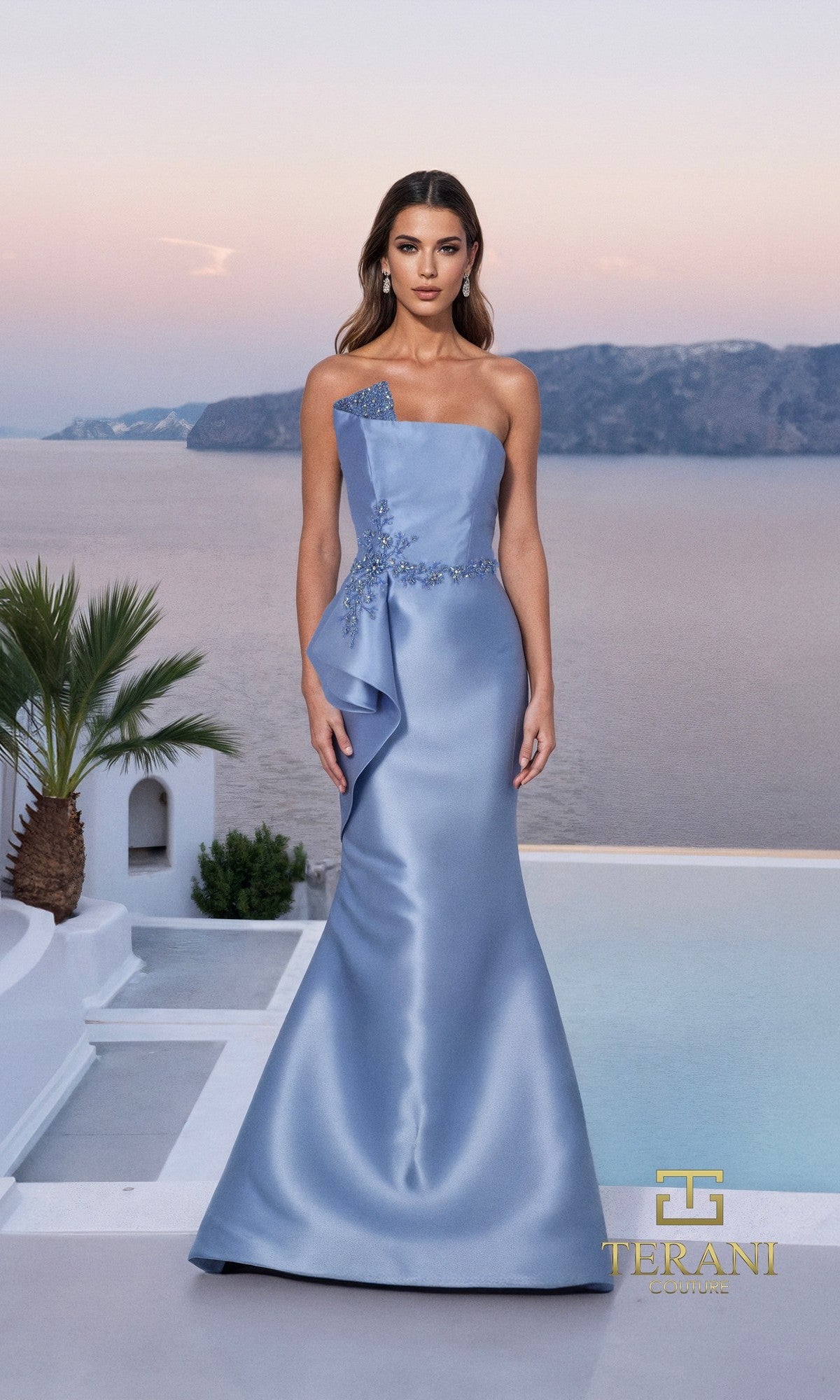 A woman in a strapless, slate Long Formal Dress: Terani 261M6462 with floral details and a mermaid silhouette stands by a pool overlooking the sea and mountains at sunset.