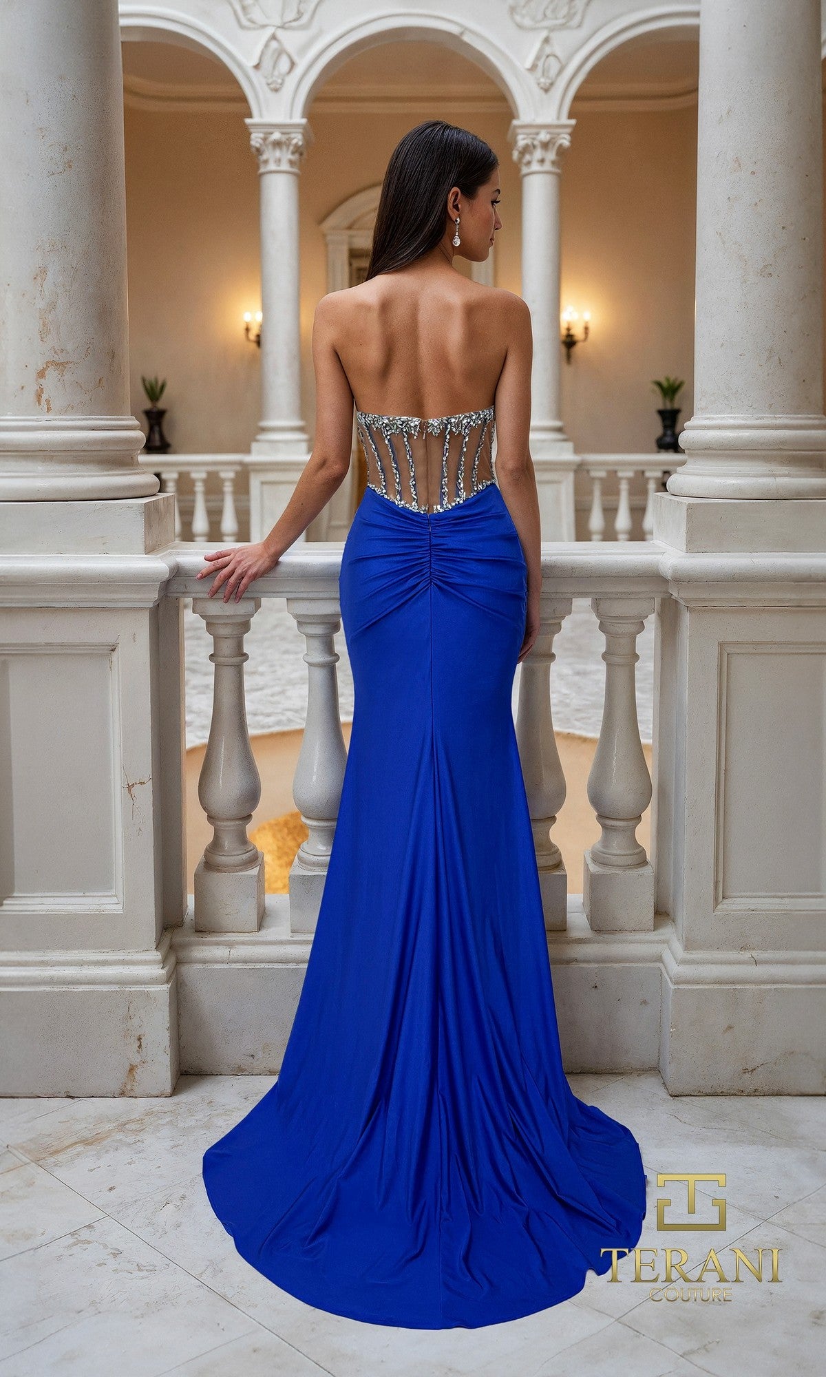 A woman stands facing away on a marble balcony, wearing the Terani 261P6115 long formal dress with a strapless royal blue design, jeweled detailing, and a sheer corset bodice.