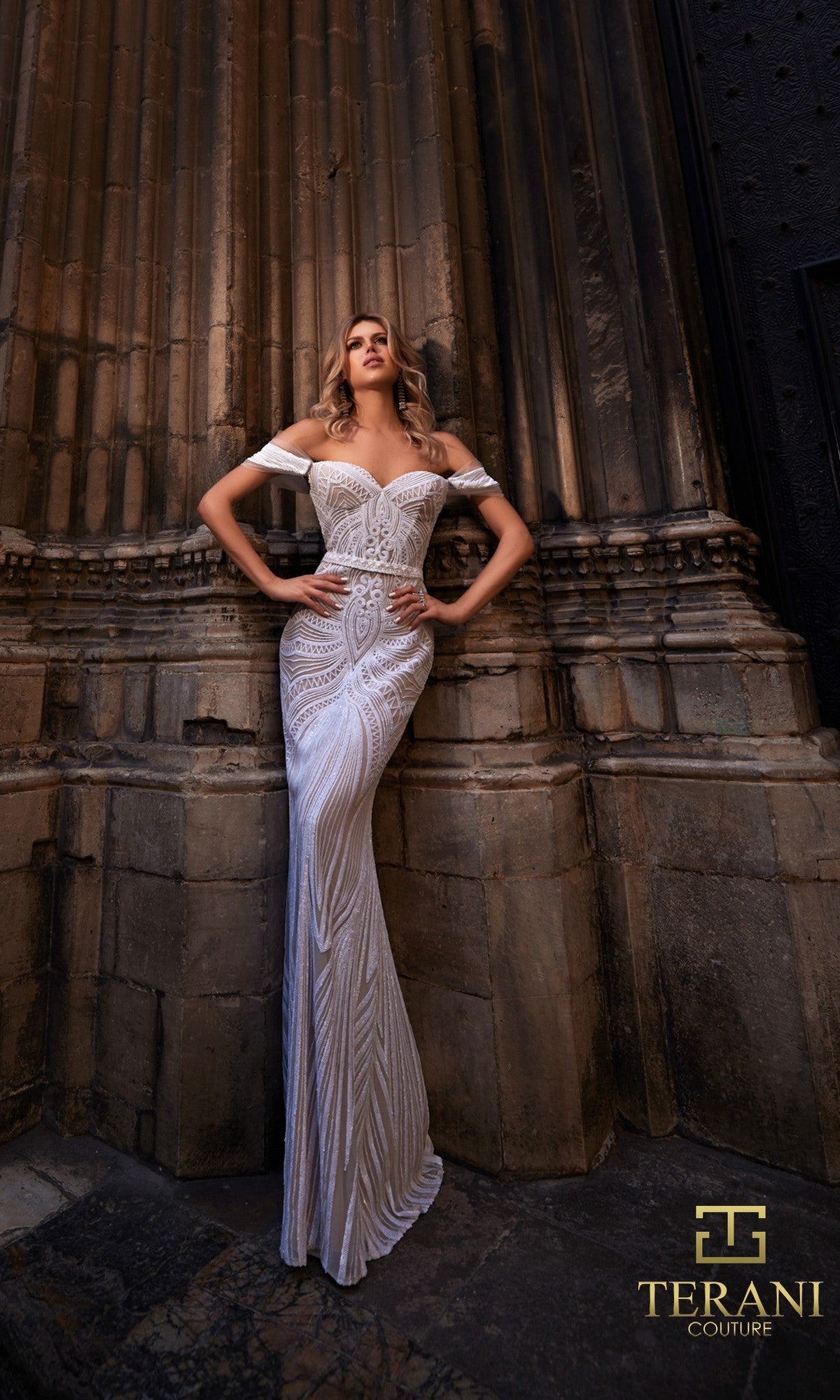 A woman poses against an ornate stone wall, wearing the Terani 261P6161 long formal dress?a white off-the-shoulder gown with intricate embroidered organza patterns. The Terani Couture logo appears in the lower right corner.