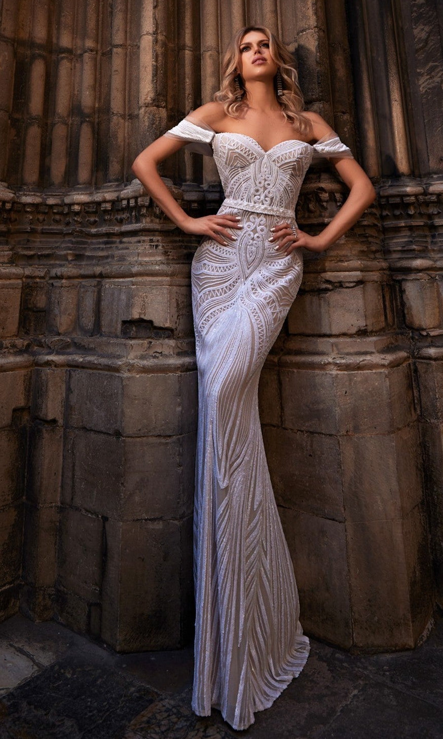 A woman in the ivory nude Terani 261P6161 long formal dress with elegant embroidered organza detail stands gracefully against a stone wall.