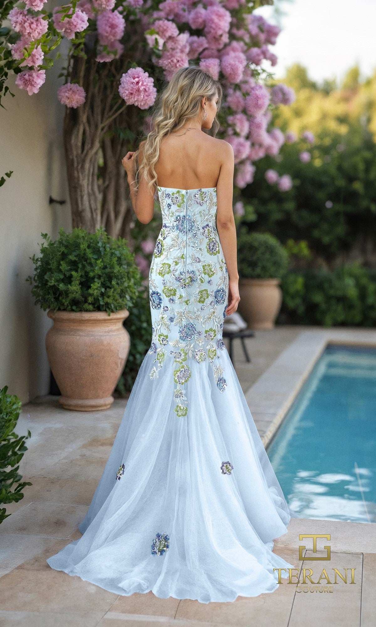 A woman in the Terani 261P6193 long formal dress, featuring a strapless tulle mermaid silhouette with floral embroidery, stands by a pool facing away, surrounded by large potted plants and pink flowers.