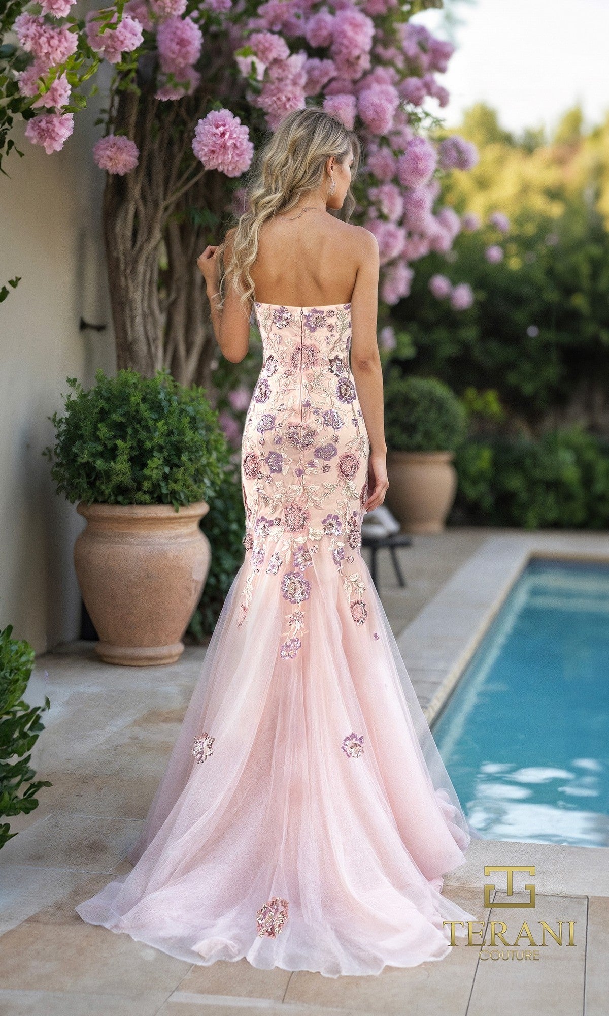 A woman with long blond hair wears the Terani 261P6193 long formal dress, a strapless pink floral mermaid gown, while standing by a pool and potted plants under blooming pink flowers.