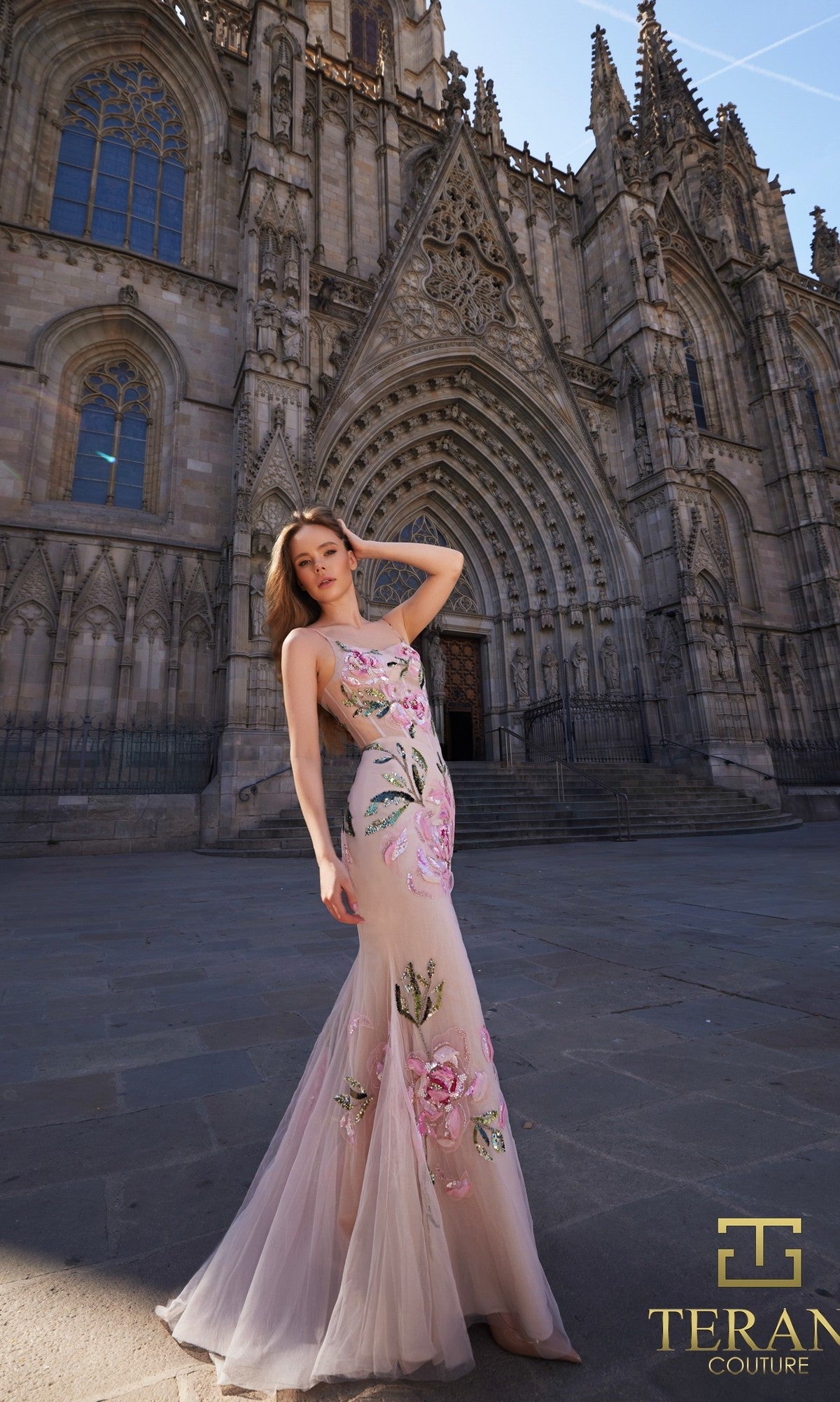 A woman in the Terani 261P6677 long formal dress, a fitted mermaid gown with floral embroidery and beading, poses before a grand Gothic cathedral. The Terani Couture logo is displayed in the corner.