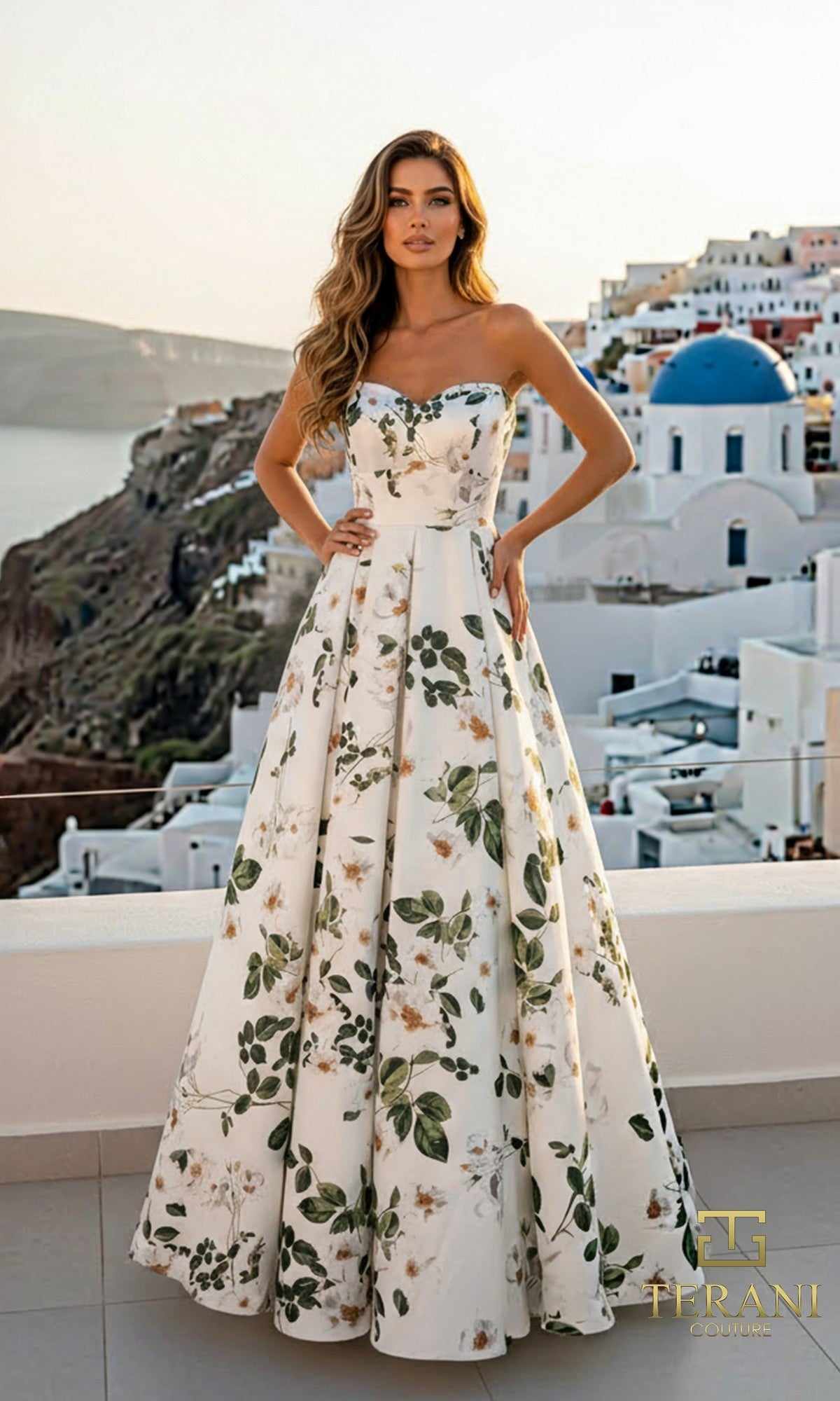 A woman in a strapless, floor-length Terani 261P6705 green multi floral satin dress with an a-line skirt stands on a terrace with white buildings and blue domes in the background.
